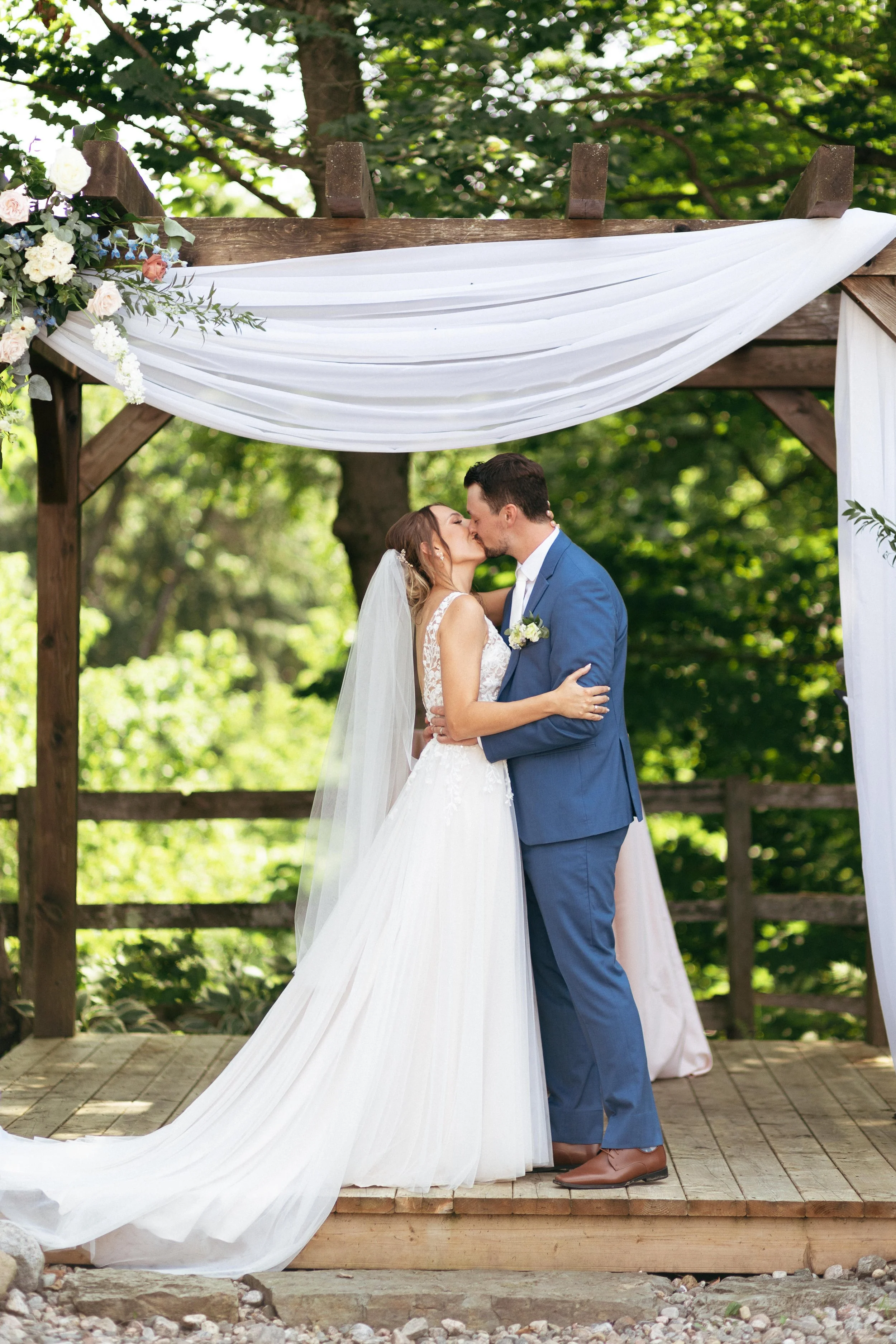 A bride and groom are kissing under a wooden wedding arch draped with white fabric and decorated with flowers, outdoors surrounded by green trees.
