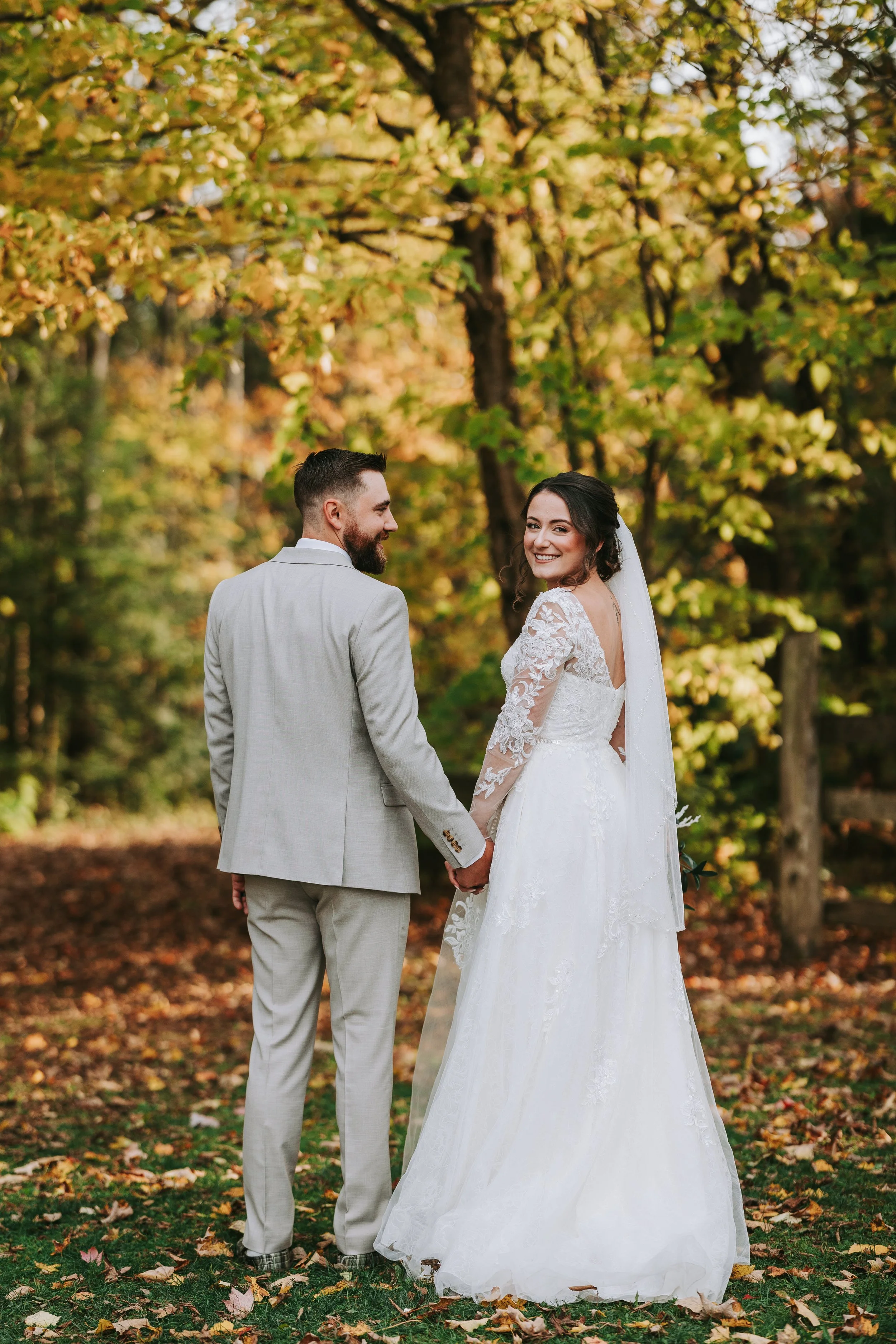 A bride and groom holding hands and smiling in an outdoor setting with autumn leaves and trees.