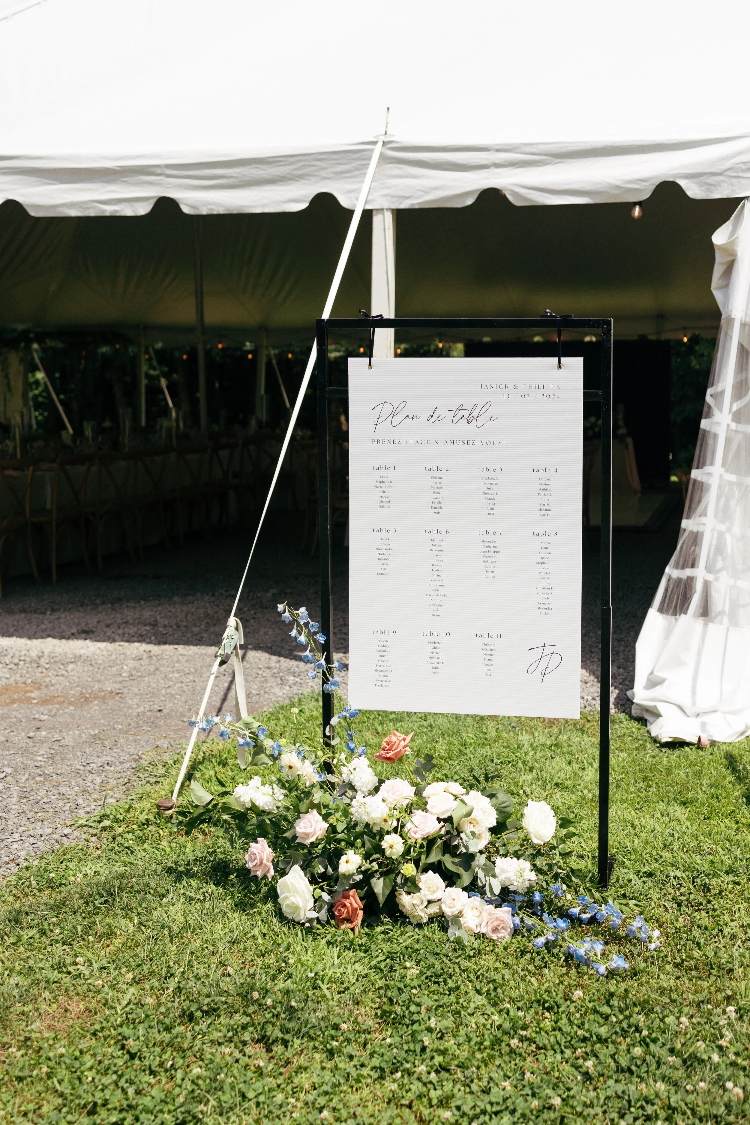 Sign displaying a wedding seating chart with floral arrangements in front, set up outdoors under a white tent.