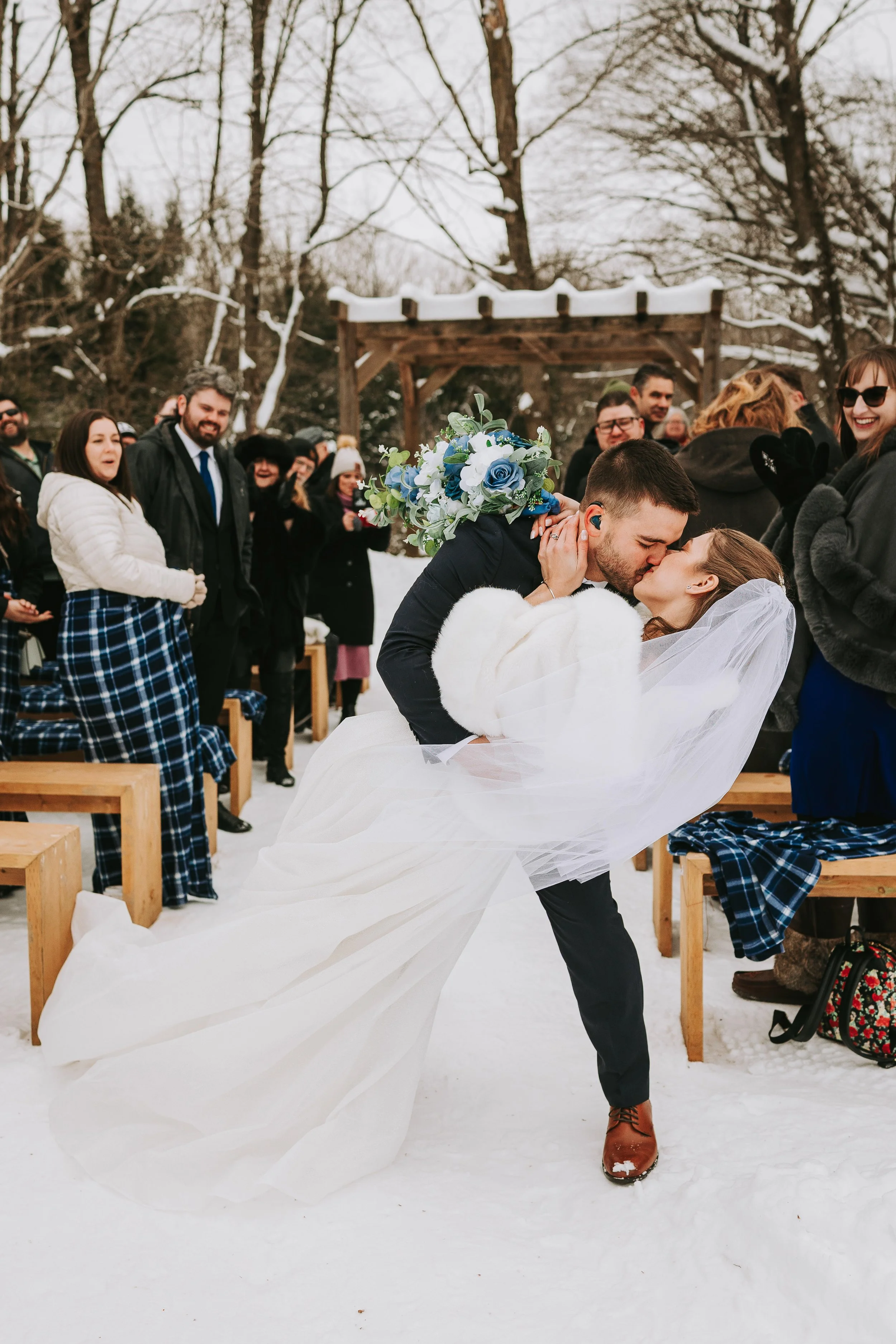 A newlywed couple sharing a kiss during an outdoor winter wedding, surrounded by friends and family in a snow-covered setting.