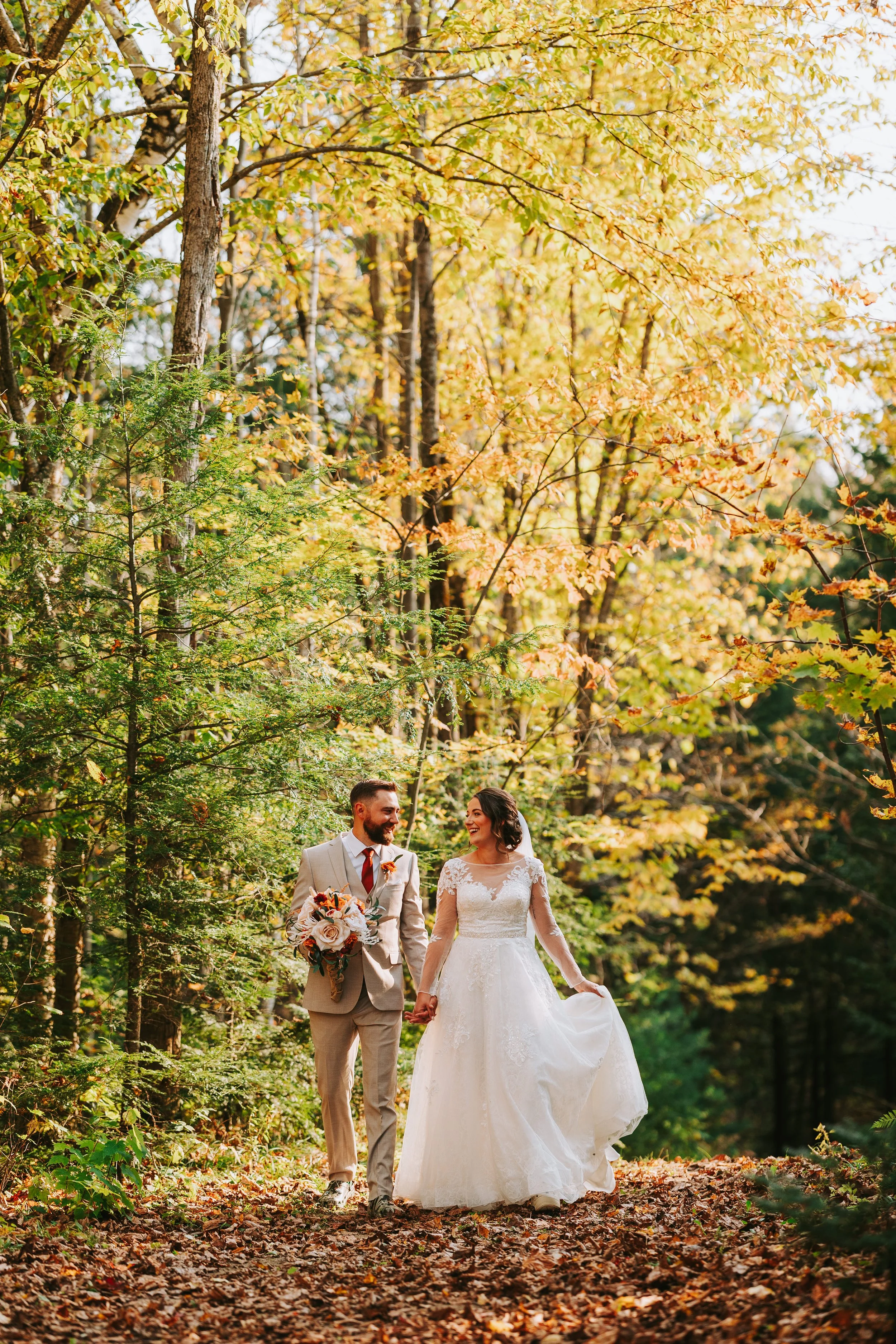 A newlywed couple holding hands and smiling while walking through a forest with autumn leaves.