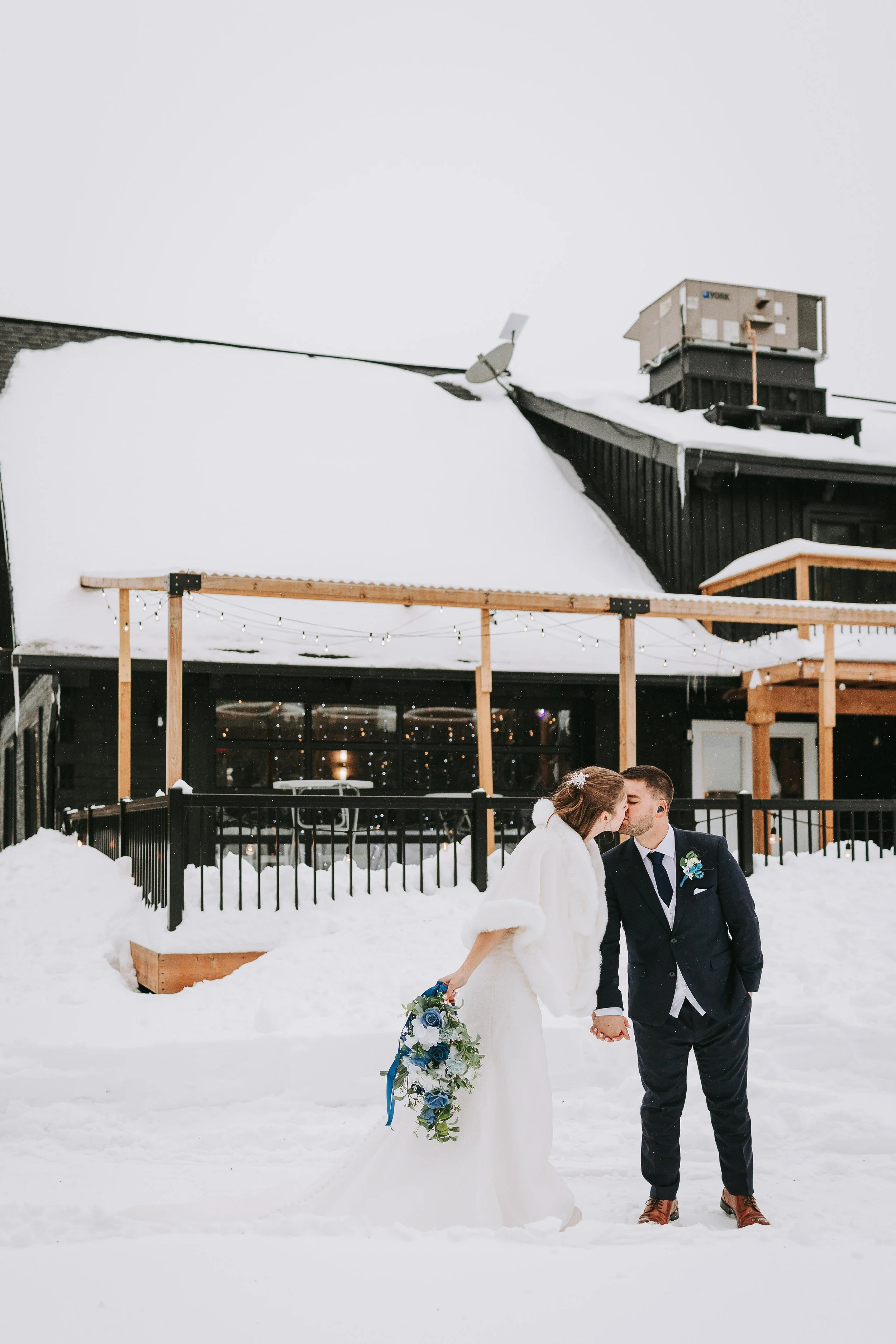 A bride and groom share a kiss outdoors in the snow on their wedding day, with snow-covered ground and a black building in the background.