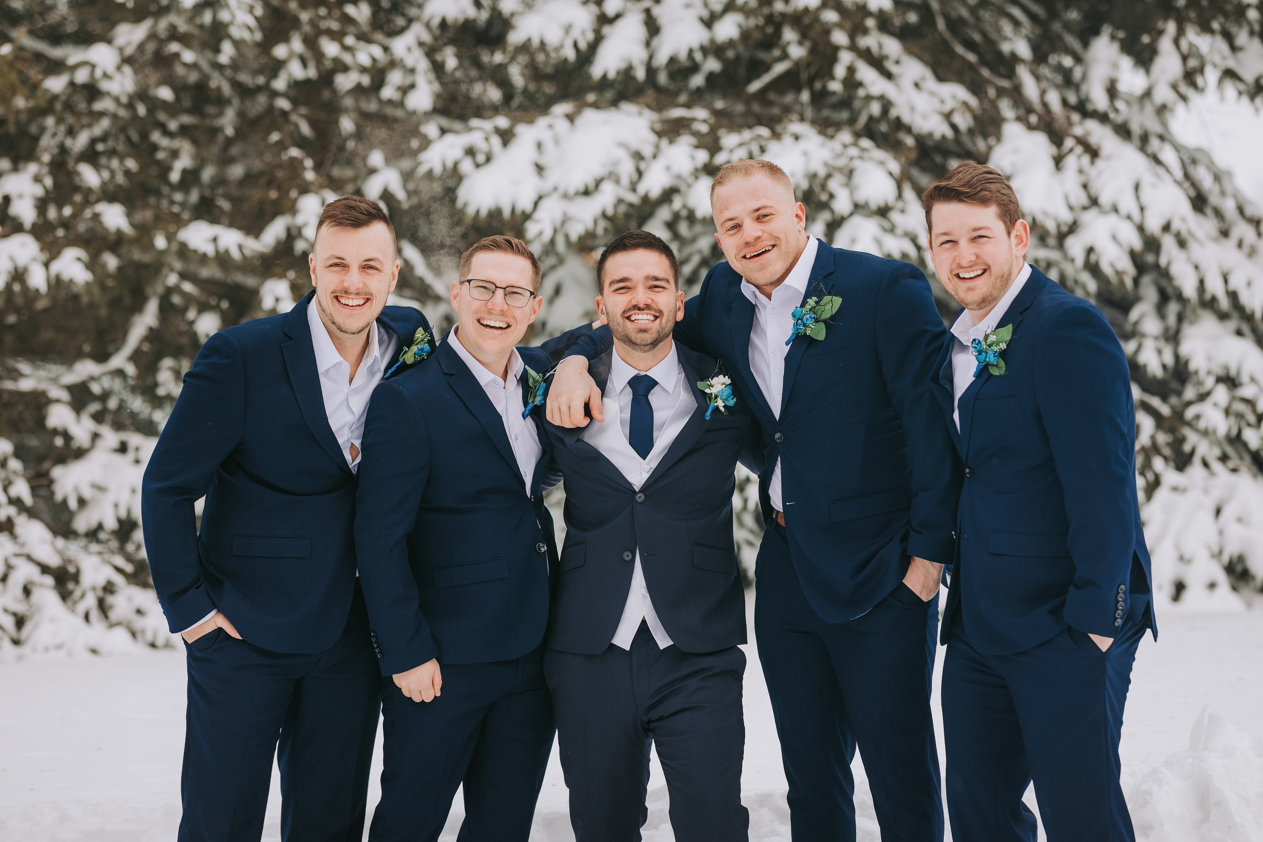 Group of five men in navy suits standing outdoors in snow, smiling for a photo, with snow-covered trees in the background.