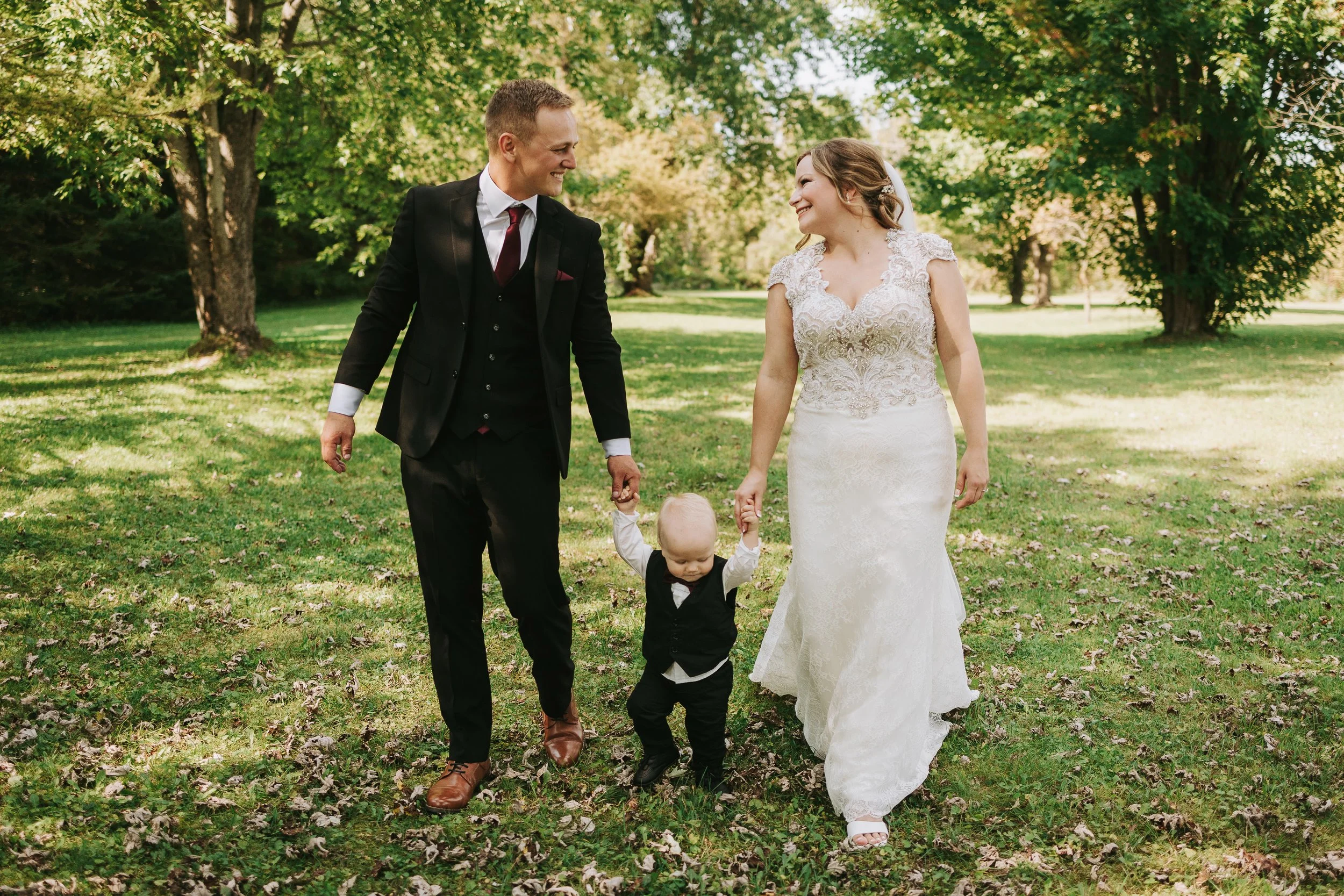A newlywed couple walking hand-in-hand with a young child in a park filled with trees and green grass.
