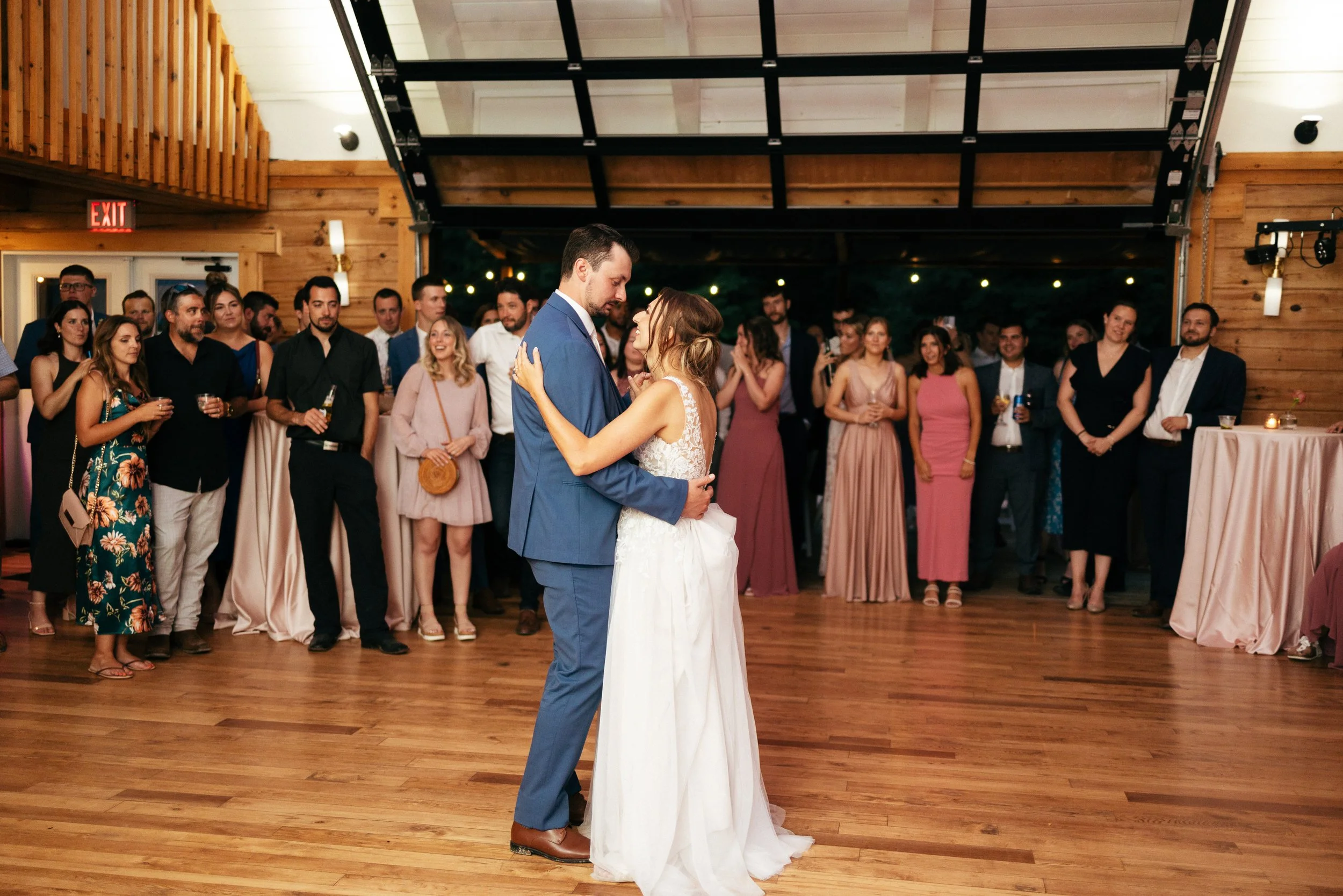 A bride and groom share their first dance at a wedding reception, surrounded by guests in a wedding venue with wooden walls and floors.
