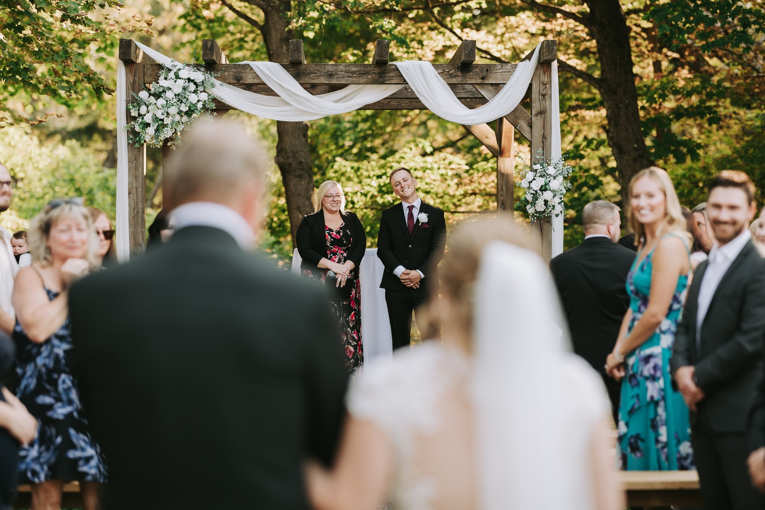 A wedding ceremony outdoors with a wooden arch decorated with white fabric and flowers, with the bride and groom standing and smiling in front of guests.