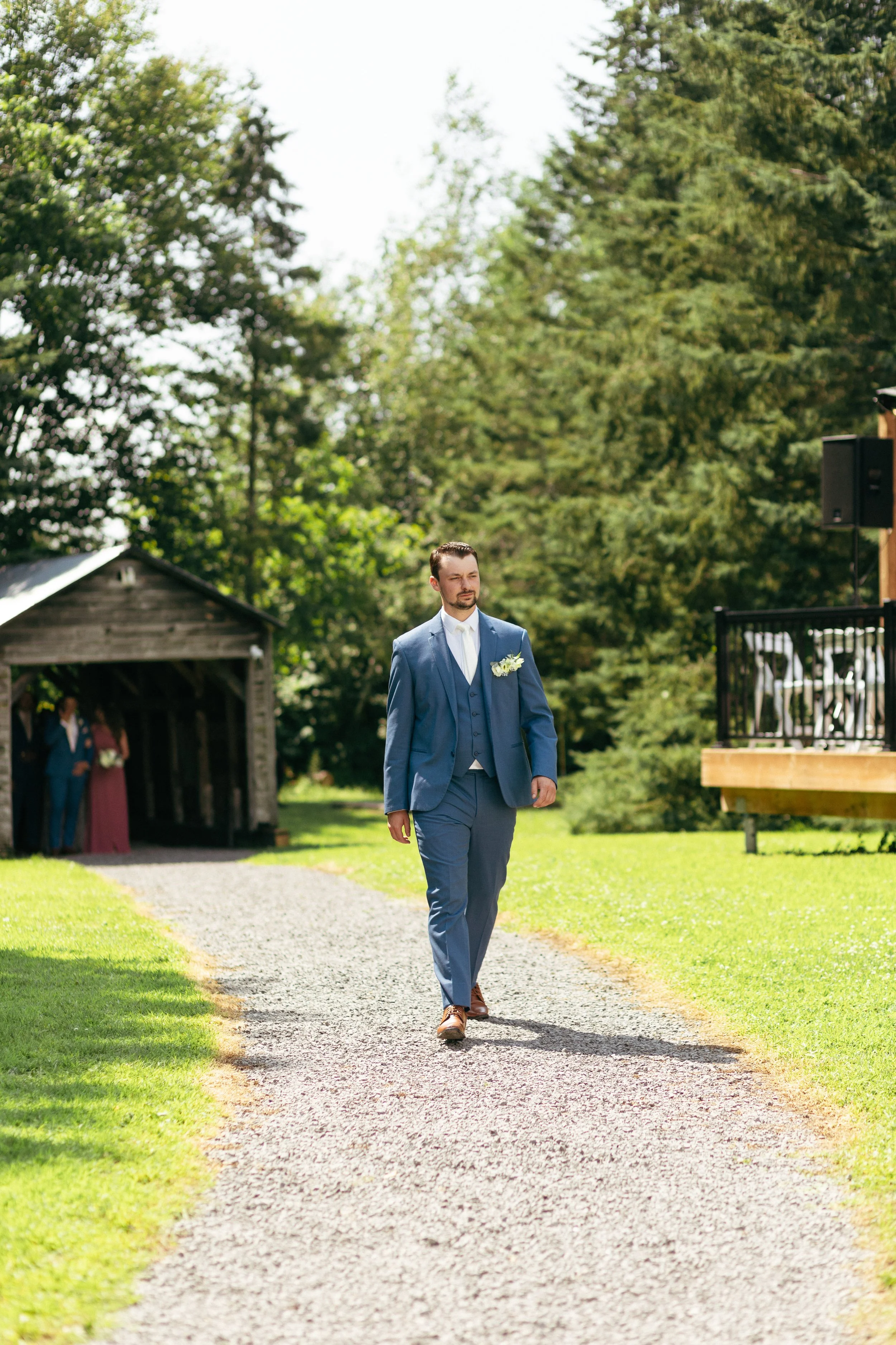 A man in a blue suit walking on a gravel path outdoors during a wedding ceremony.