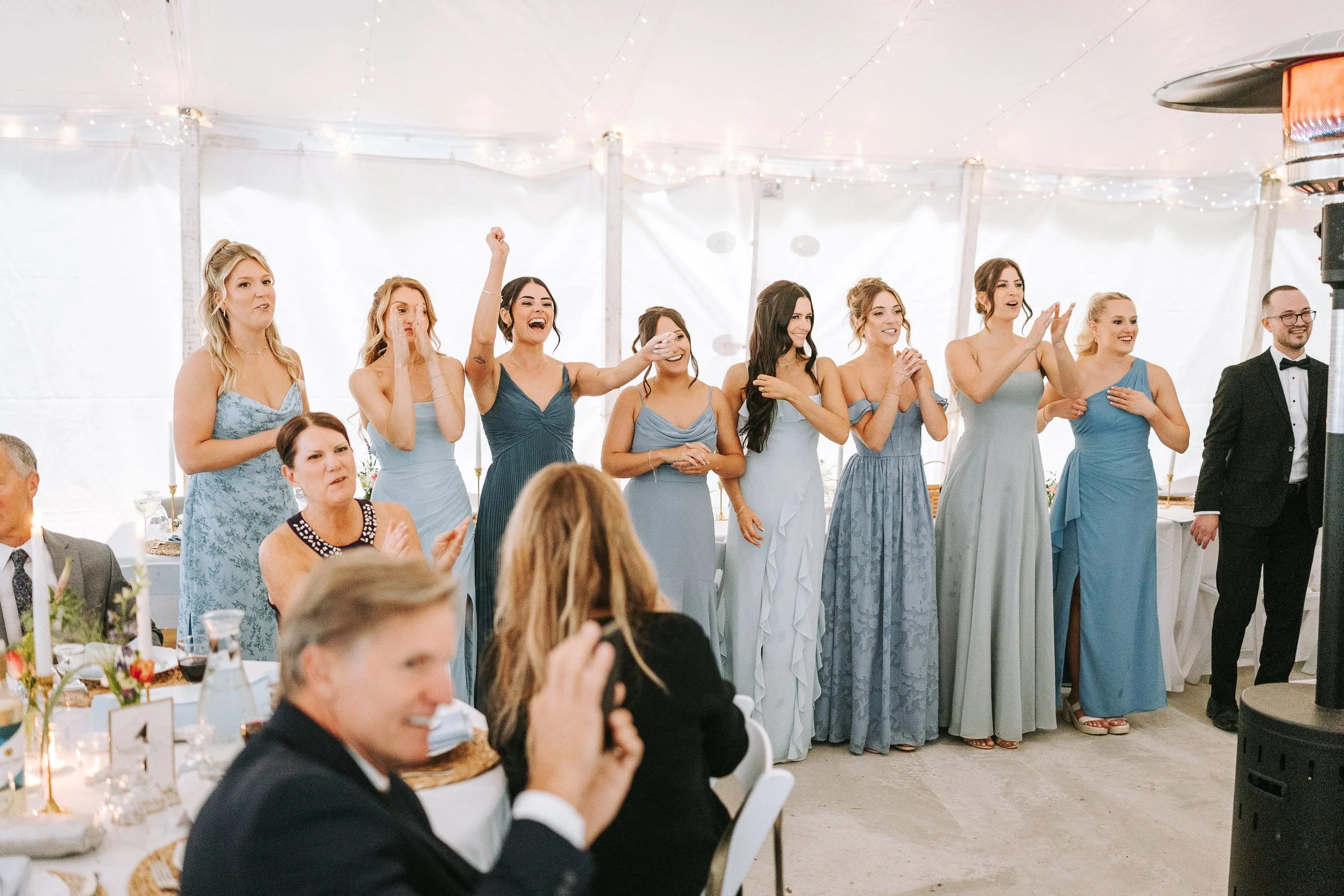 A group of women in formal dresses standing together, watching an event and clapping in a festive indoor setting with fairy lights. There is a man in a black tuxedo with a bow tie on the right.