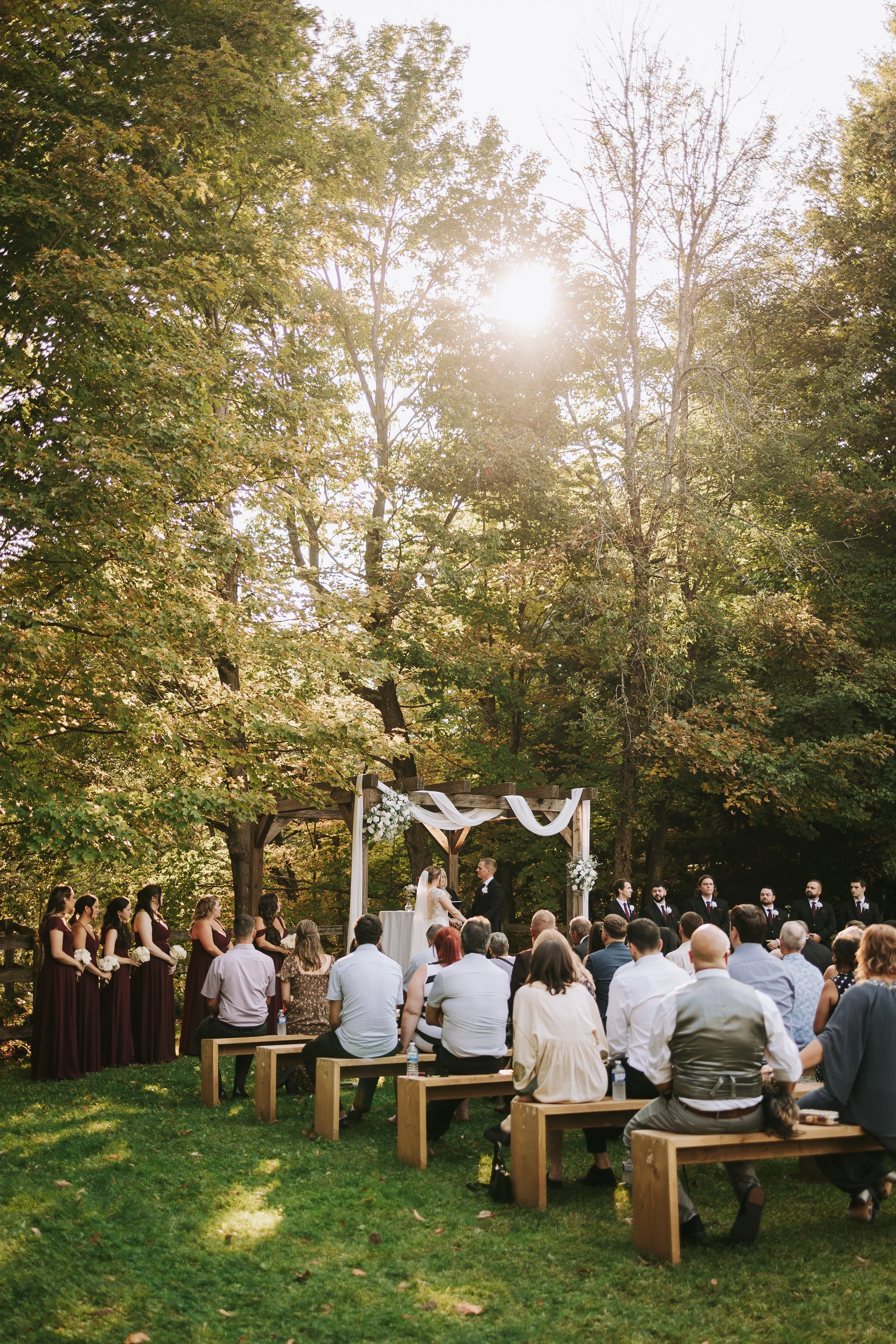 An outdoor wedding ceremony taking place in a forested area during late afternoon, with guests seated on wooden benches and bridesmaids in matching dresses standing on the left side.