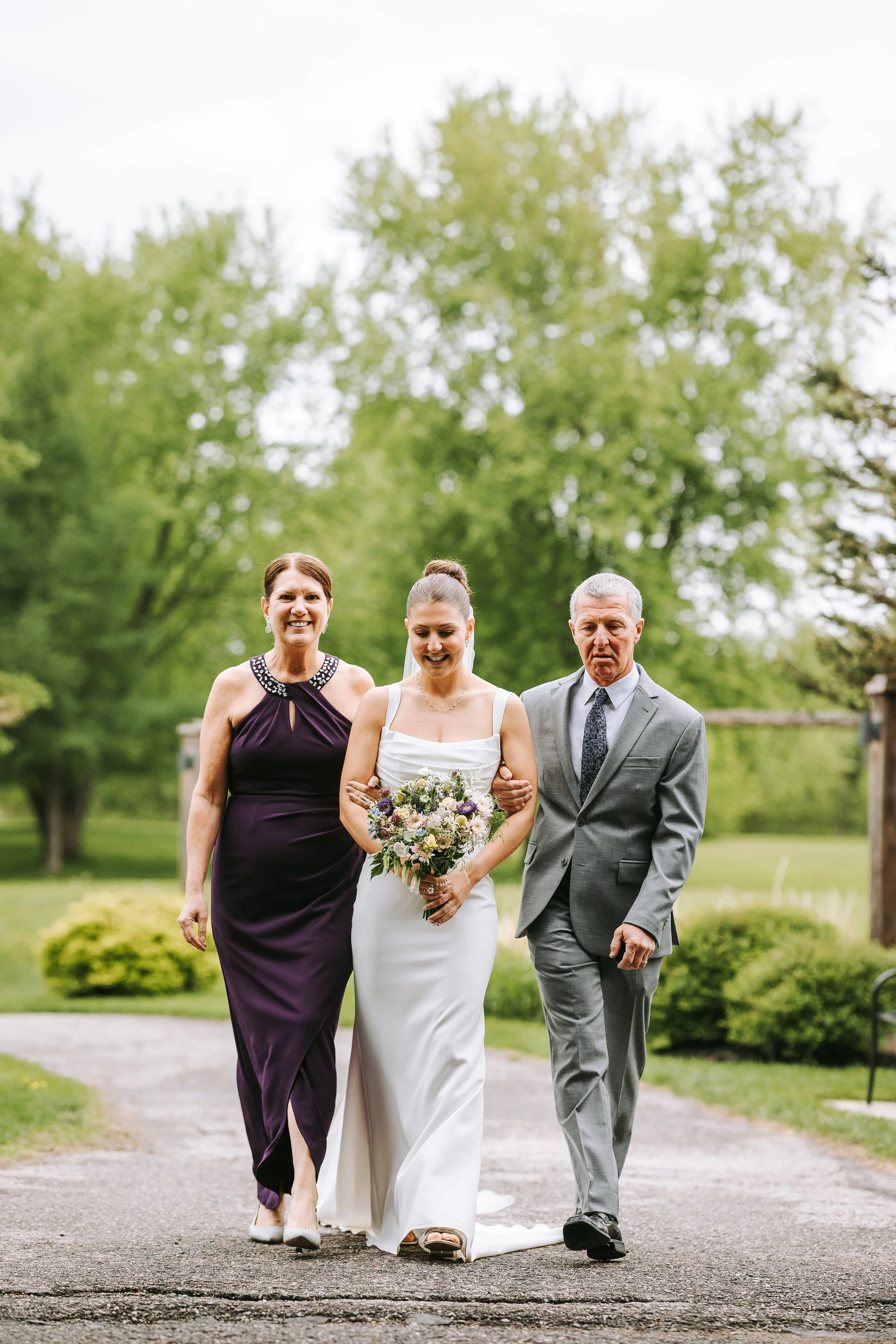 A bride walking outside with her parents, holding a bouquet of flowers, on a pathway in a park with green trees in the background.