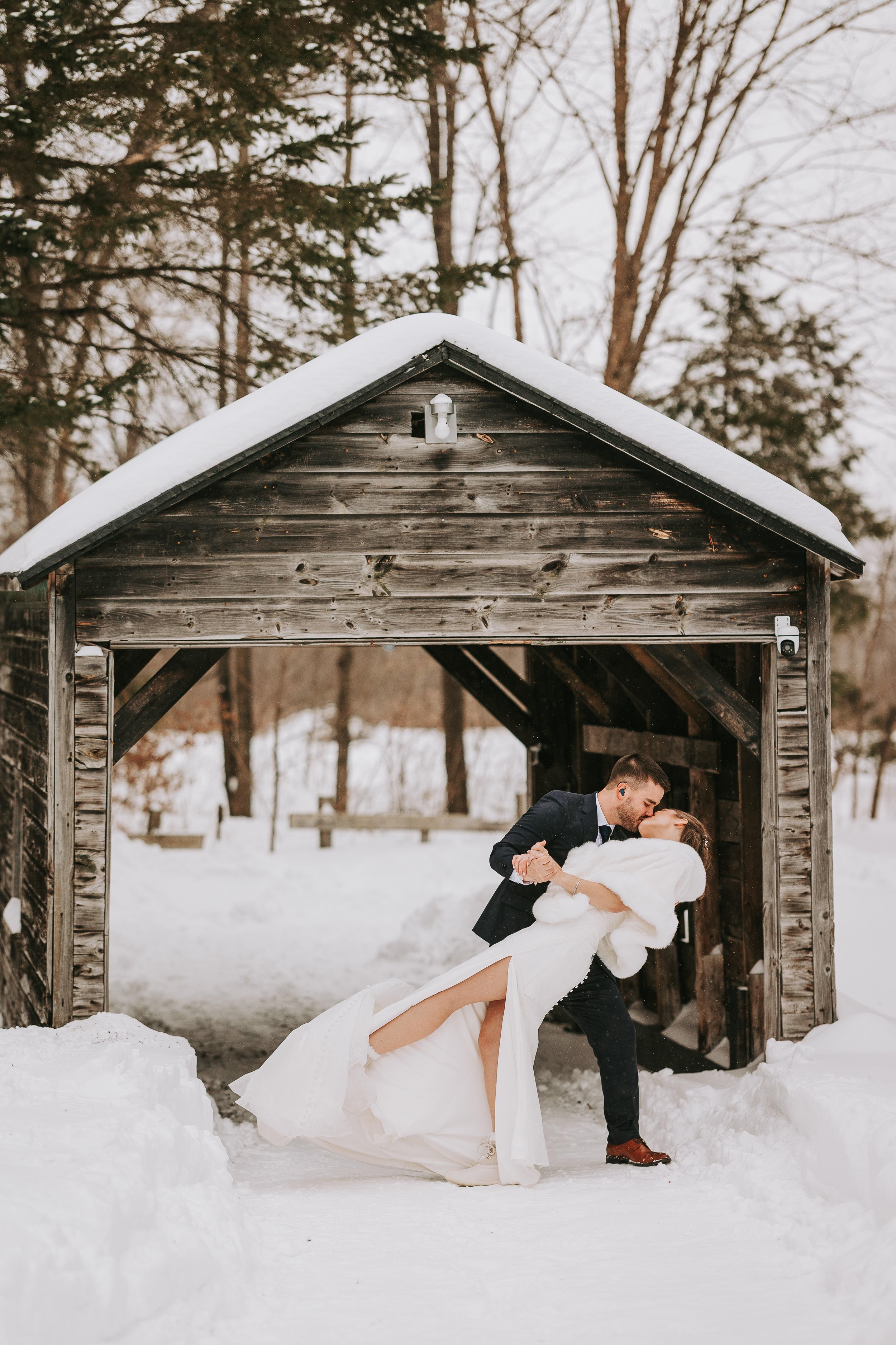 A couple in wedding attire sharing a kiss in a snowy outdoor setting, with the groom dipping the bride near a rustic wooden bridge.