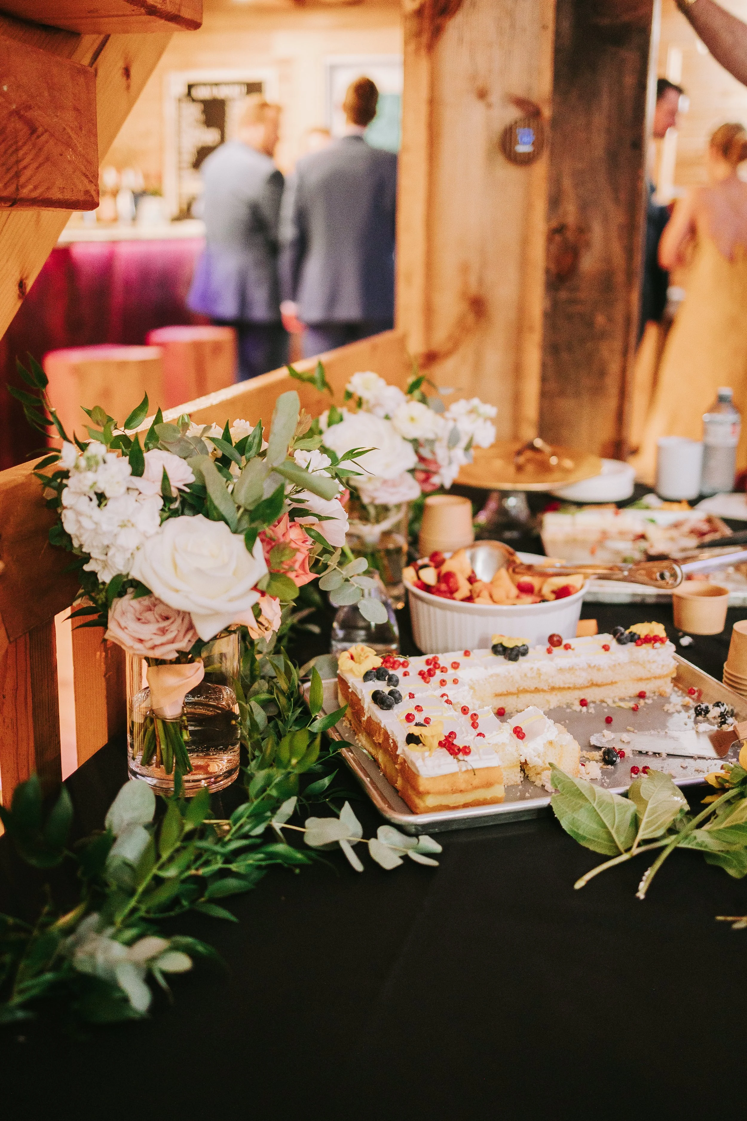 A dessert table with a partially eaten white frosted cake garnished with red and black berries, surrounded by floral arrangements with white and pink roses and greenery, set in a wedding venue with people mingling in the background.