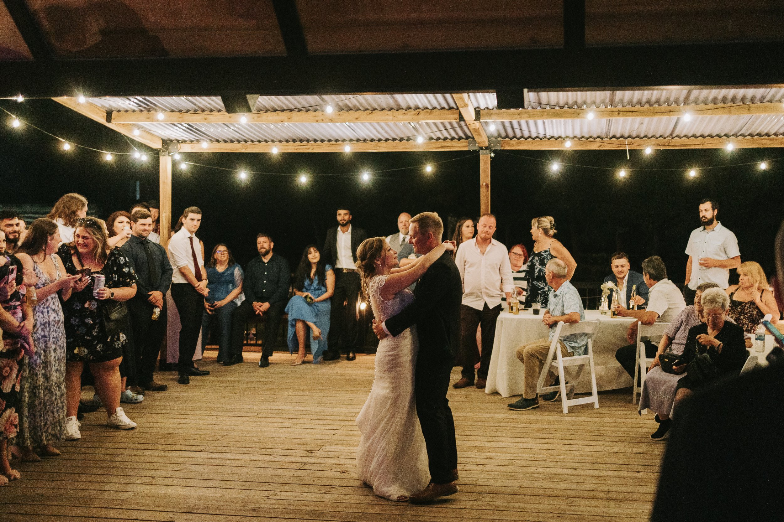 A bride and groom are dancing together at their wedding reception under a decorated outdoor pavilion at night. Guests are gathered around watching and taking photos, with string lights overhead creating a festive atmosphere.