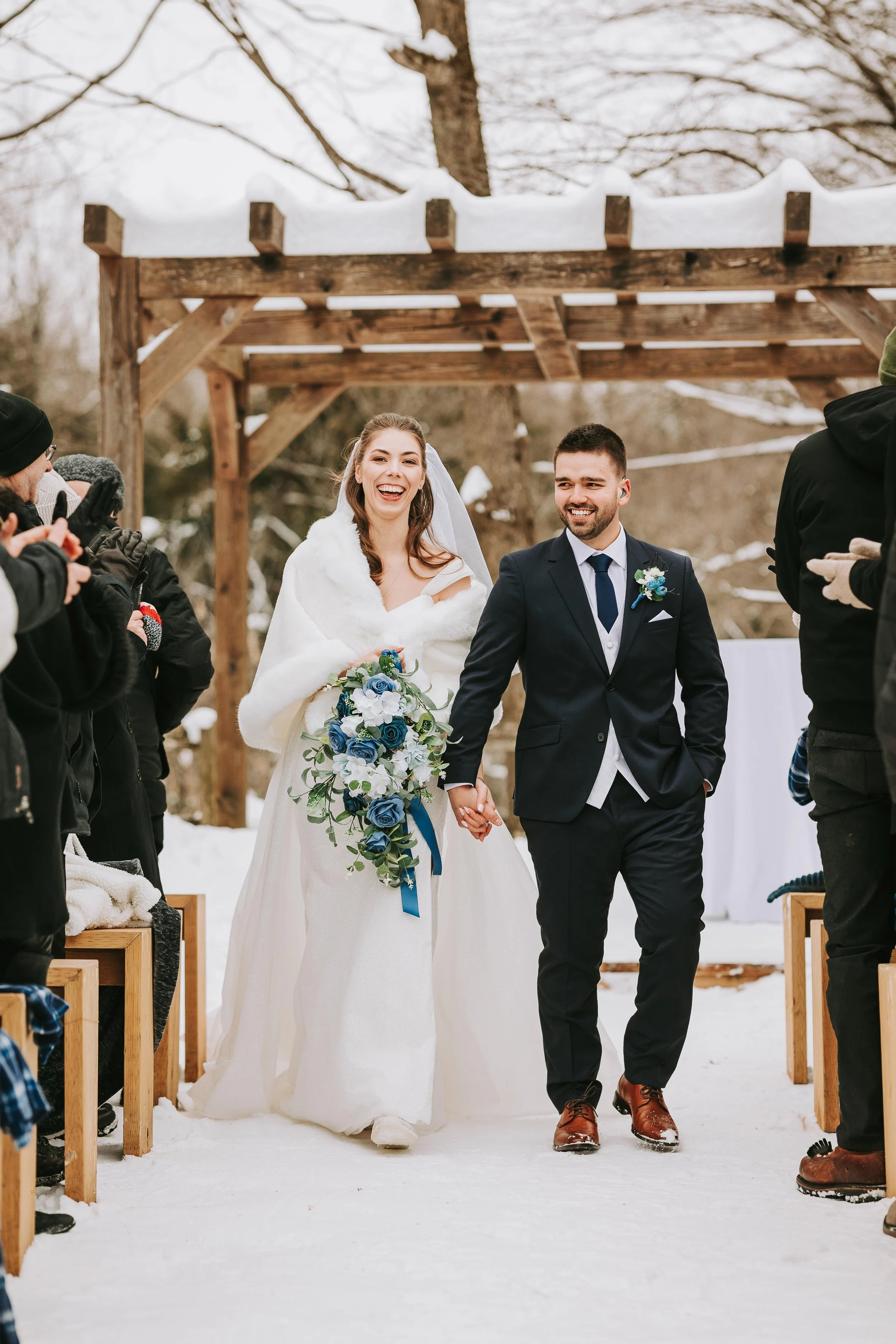 A newlywed couple walking hand-in-hand down an outdoor snowy aisle after their wedding ceremony, surrounded by friends and family.
