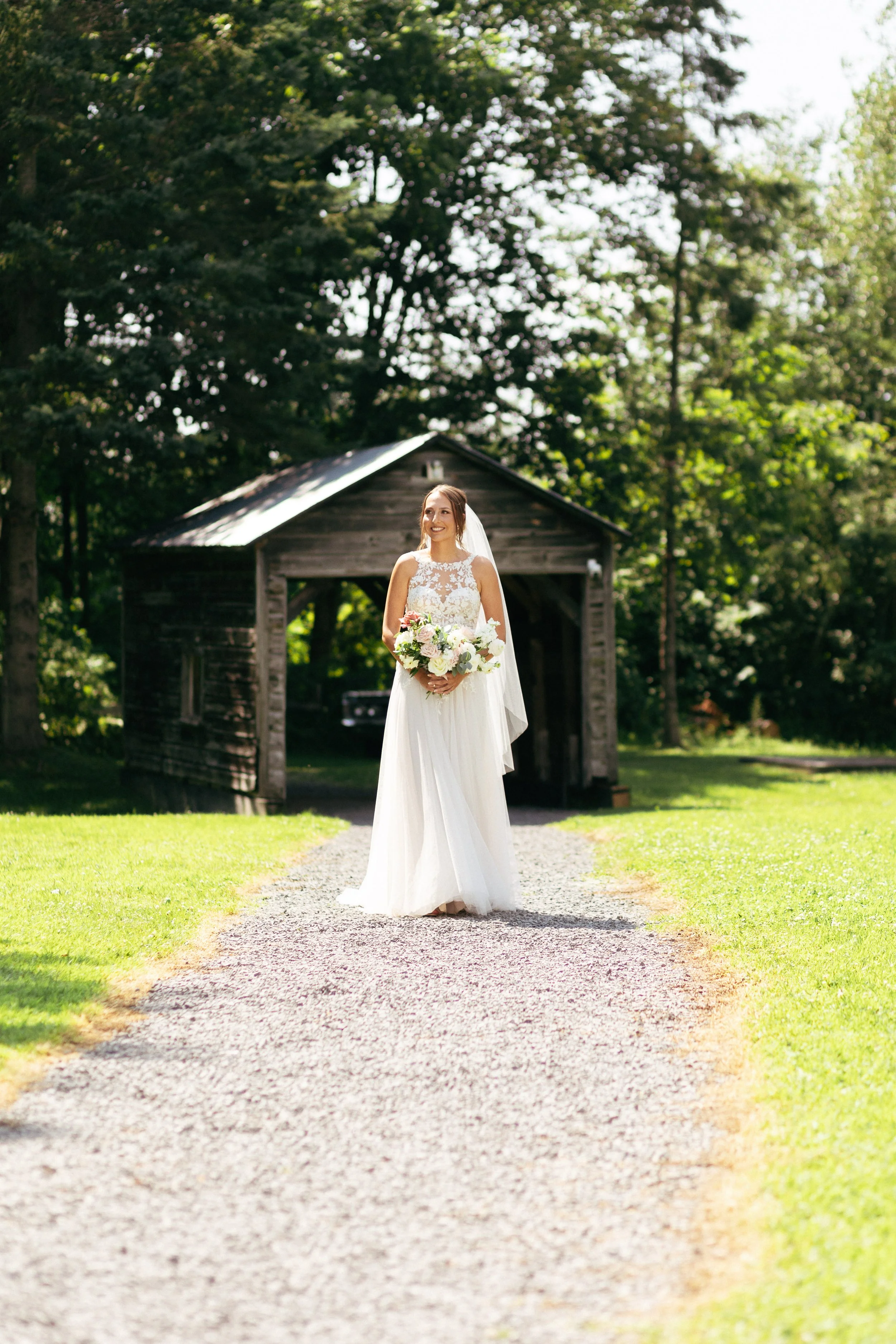 A bride in a white wedding dress holding a bouquet of flowers, standing on a gravel path with a rustic wooden barn and green trees in the background during daytime.