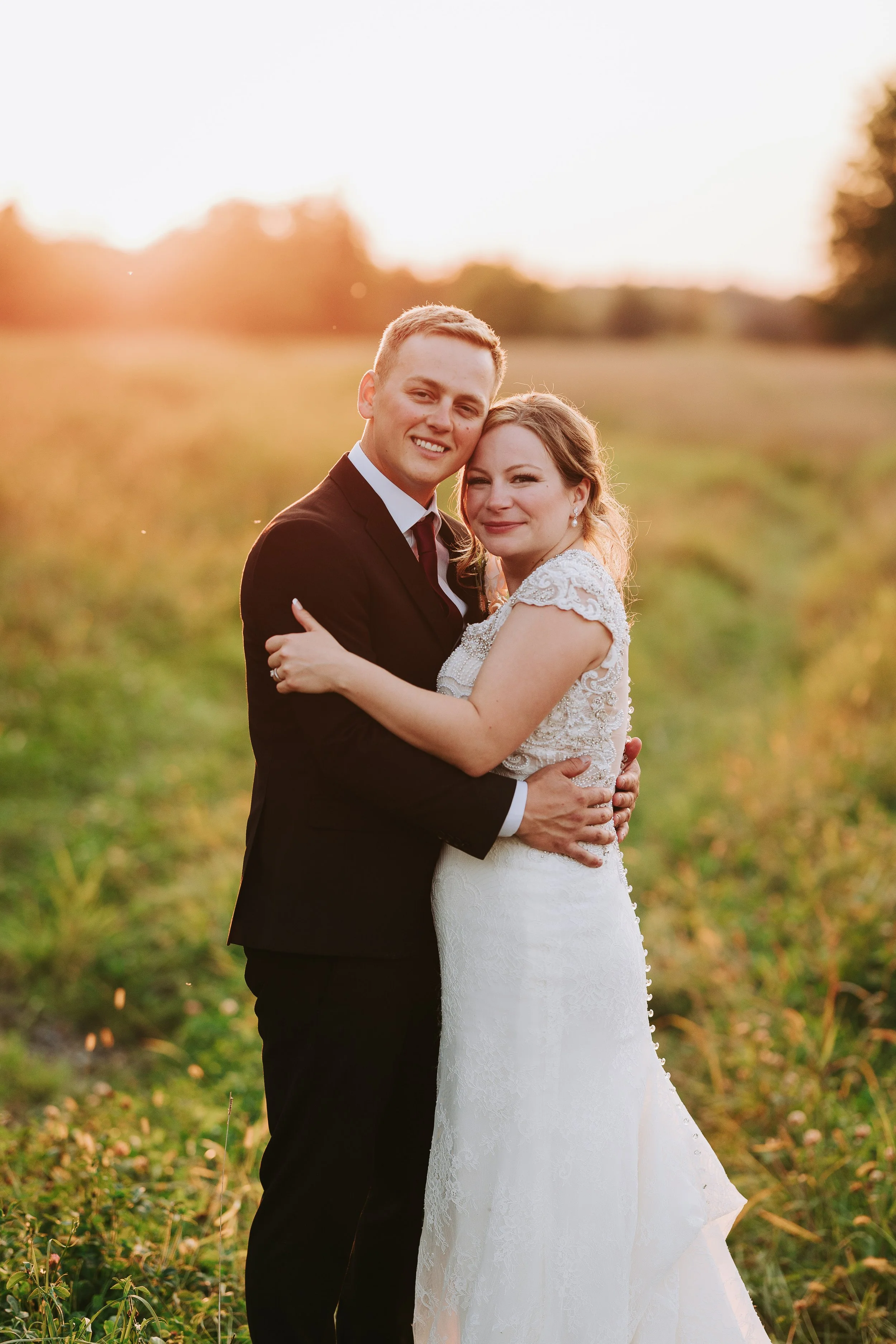 A newly married couple in wedding attire embrace outdoors at sunset, smiling at the camera, in a field with greenery and trees in the distance.