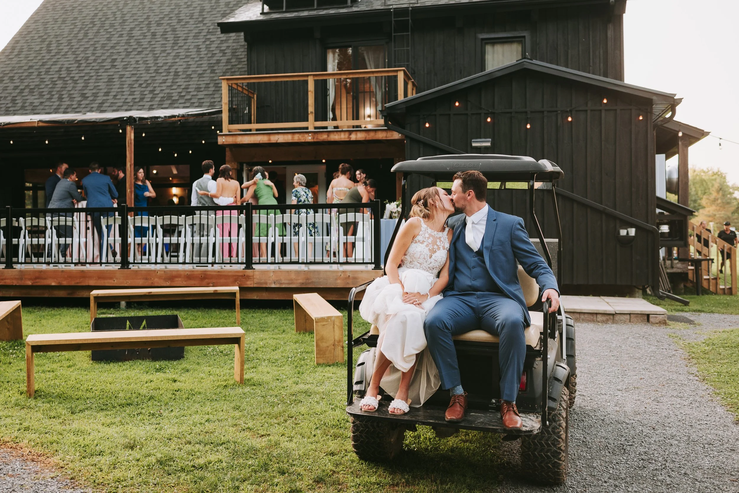 A newlywed couple sharing a kiss while sitting on a golf cart in front of a Ottawa wedding venue with wedding guests dancing and socializing on the patio in the background.