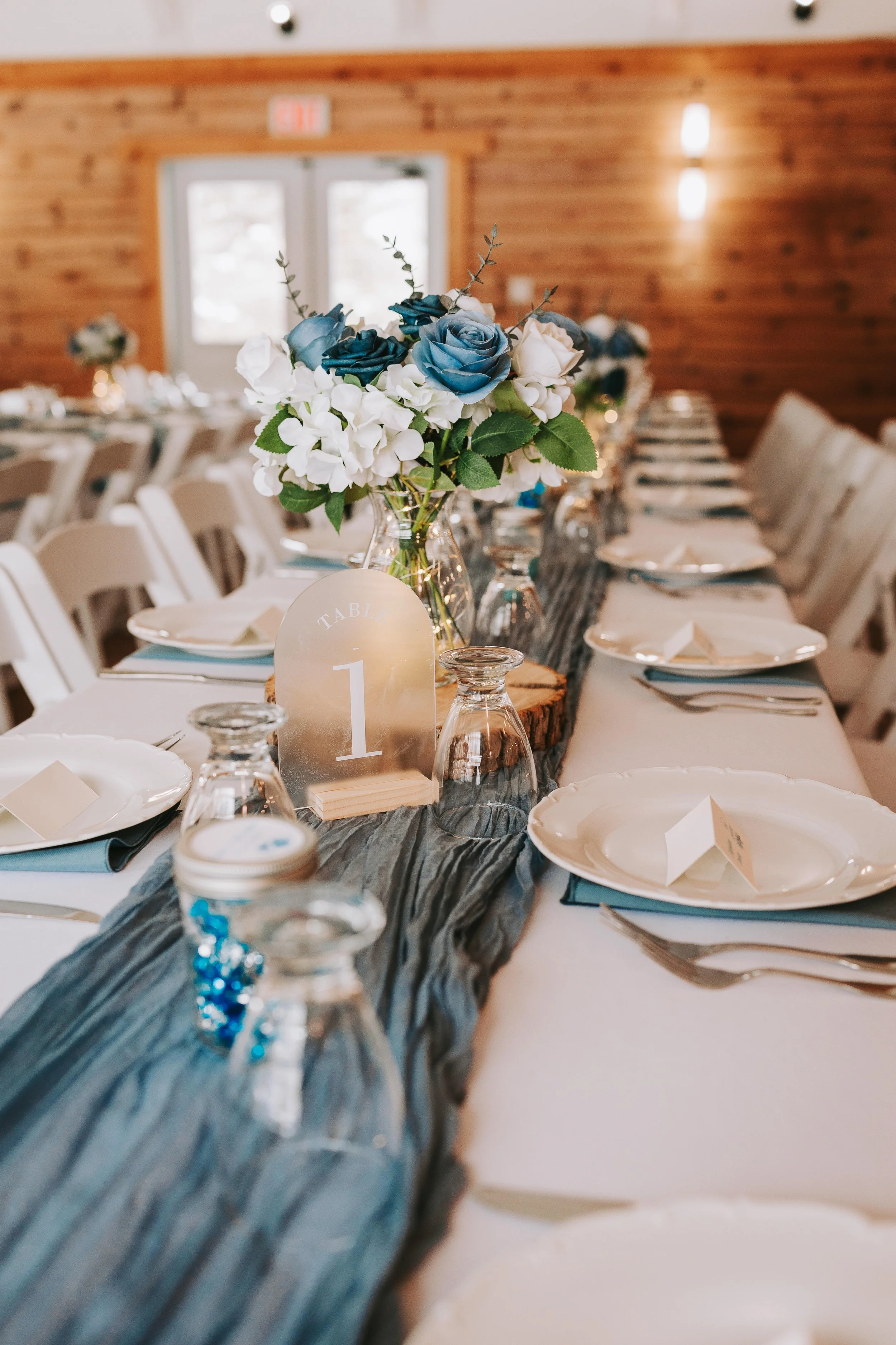 Table setting at a wedding reception with a long table, white plates, utensils, water glasses, a gray cloth table runner, and a floral centerpiece with blue and white flowers in a glass vase, with a wooden wall background.
