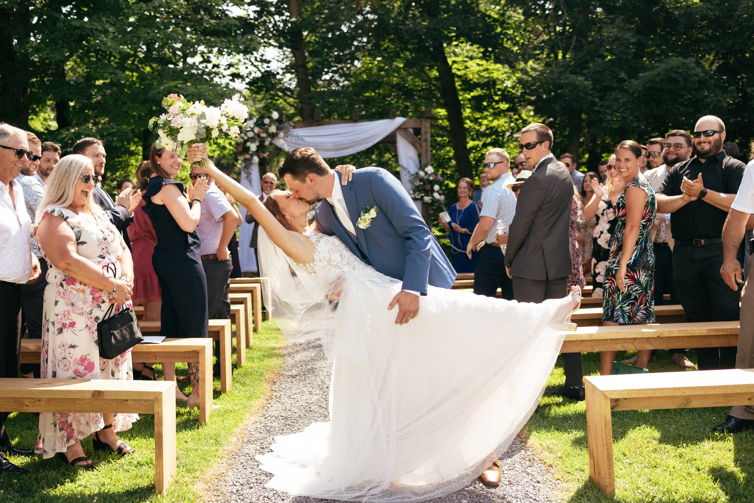 A bride and groom kiss during their outdoor wedding ceremony, with the groom dipping the bride as she holds a bouquet high above her head, surrounded by cheering guests on a sunny day in a green, wooded setting.