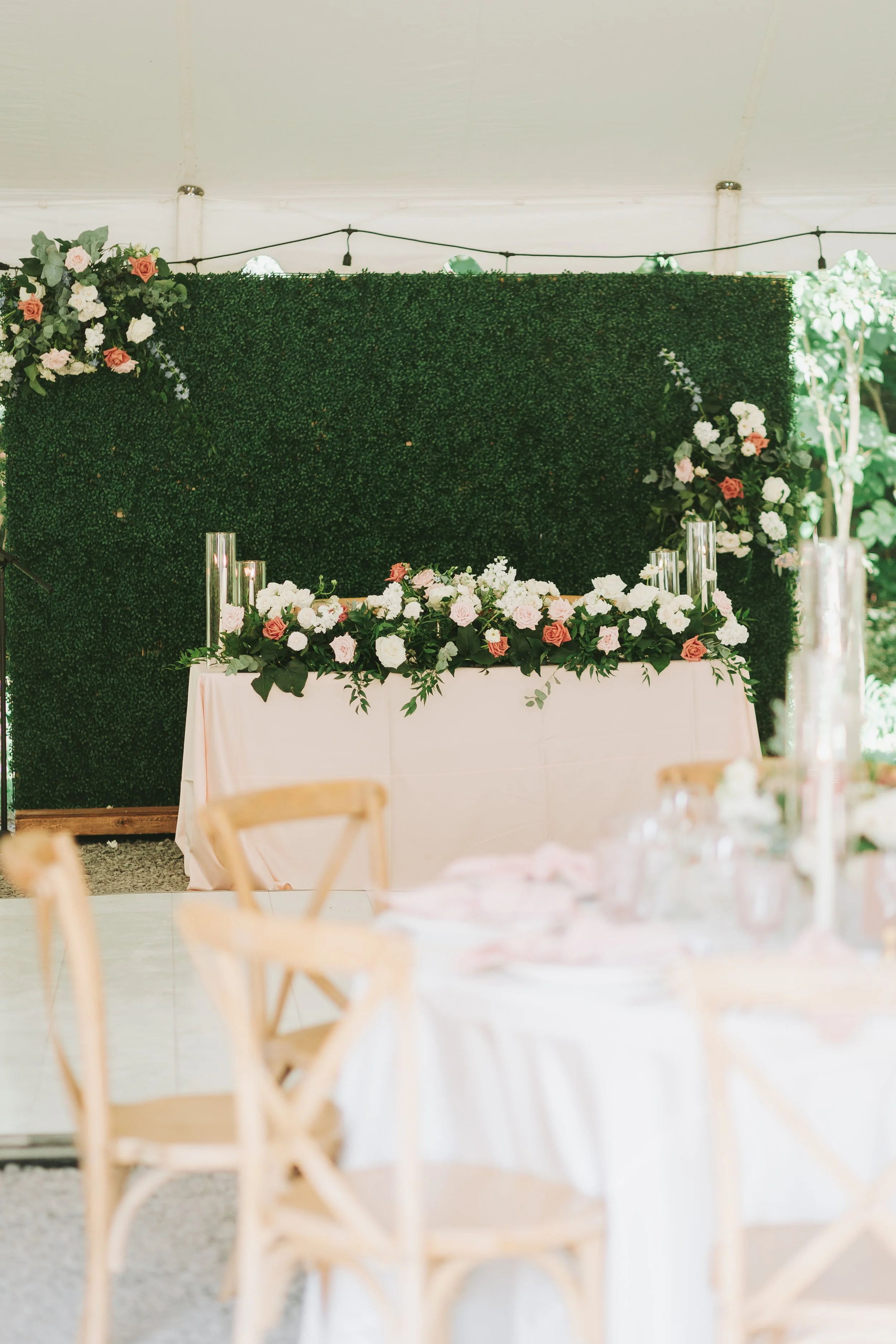 Decorated sweetheart table with pink and white flowers, green foliage, and tall glass candle holders in front of a green hedge backdrop, set up for a wedding reception in a tent.