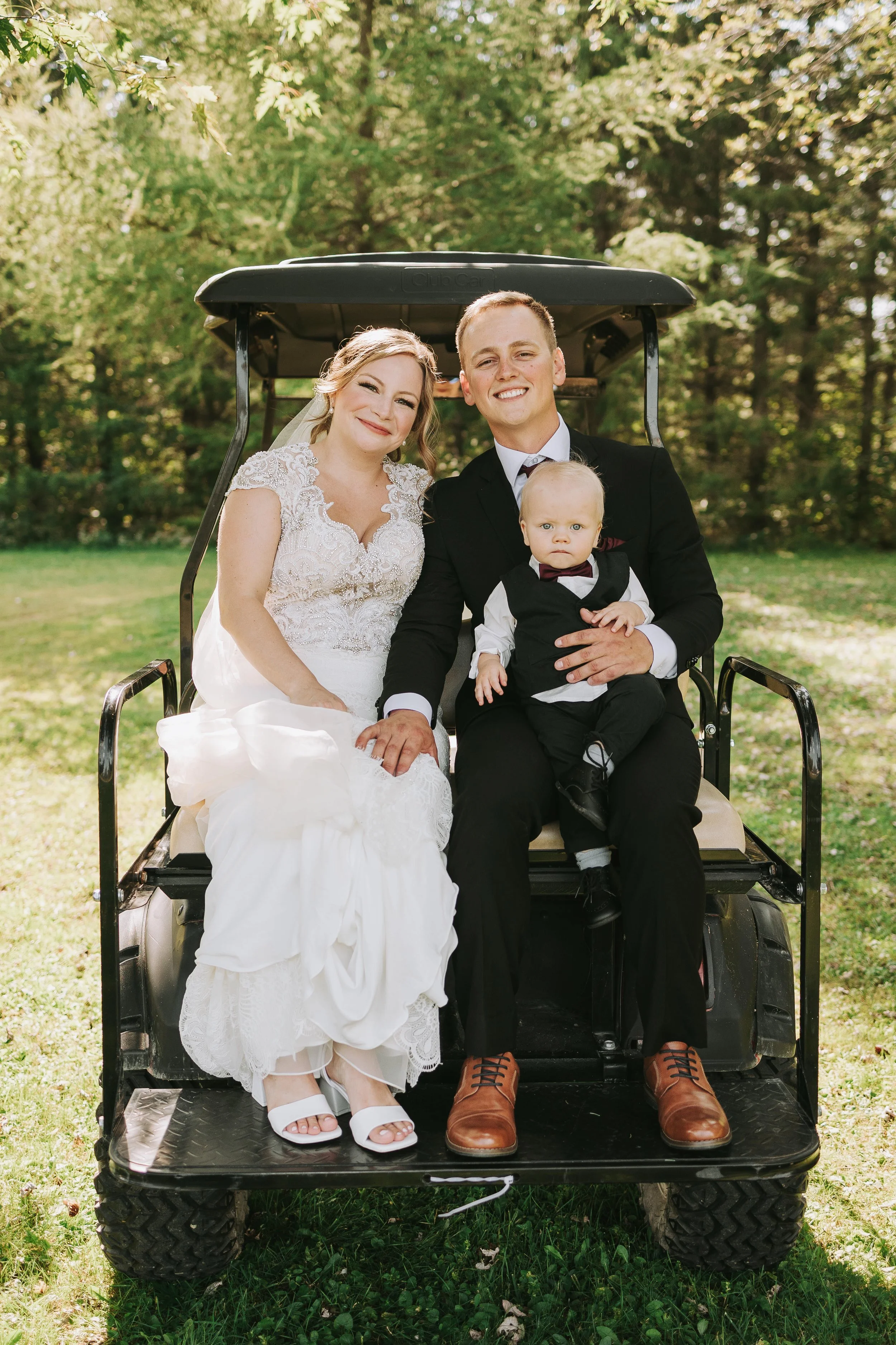 A newlywed couple with a young child on a golf cart outdoors, surrounded by trees on a sunny day.