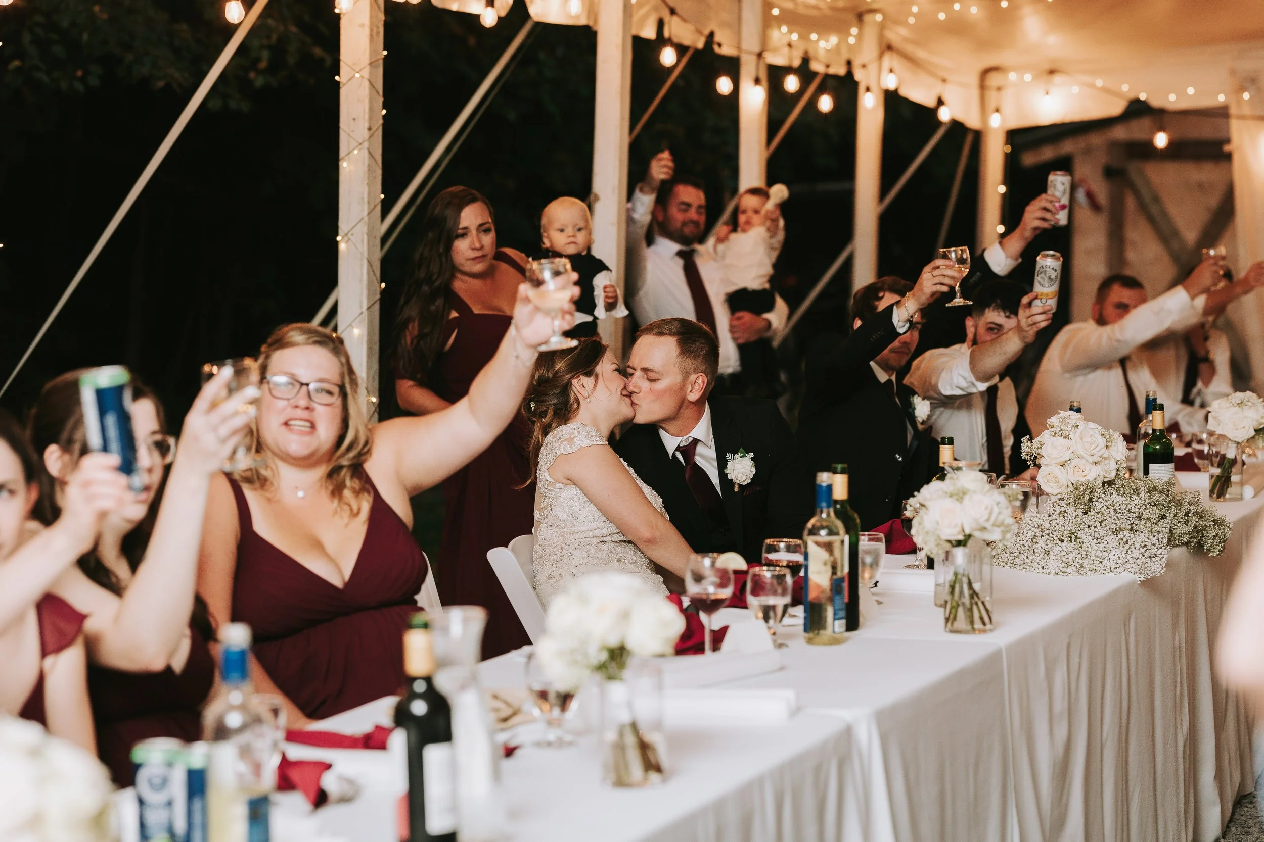 A wedding celebration with guests raising their drinks in a toast, a bride and groom kissing at the center, and people sitting and standing under a decorated tent with string lights.