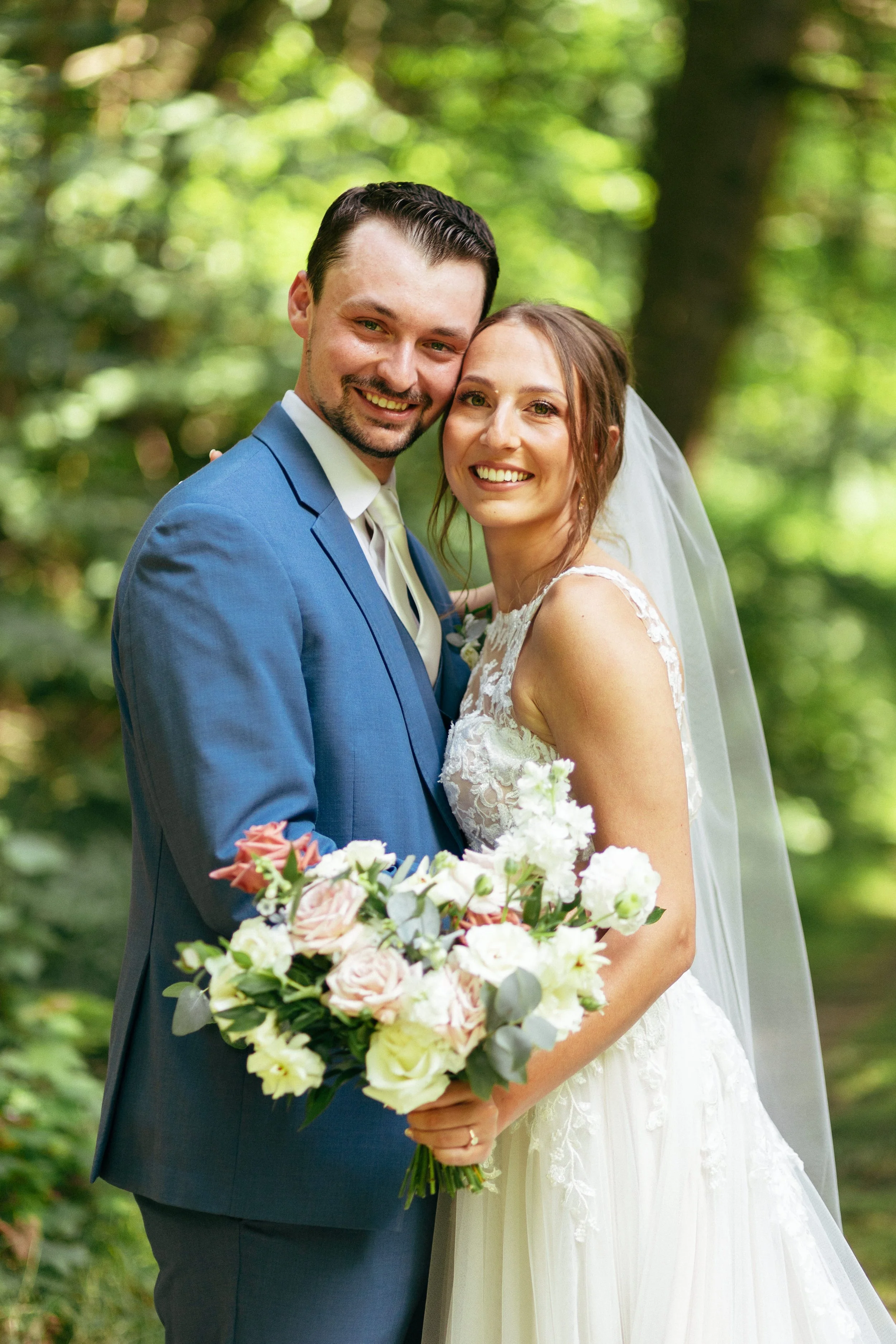 A newlywed couple is smiling and posing outdoors in a lush green forest. The bride is wearing a white lace wedding dress and a veil, holding a bouquet of pastel-colored flowers. The groom is dressed in a blue suit with a white shirt and tie, and has 