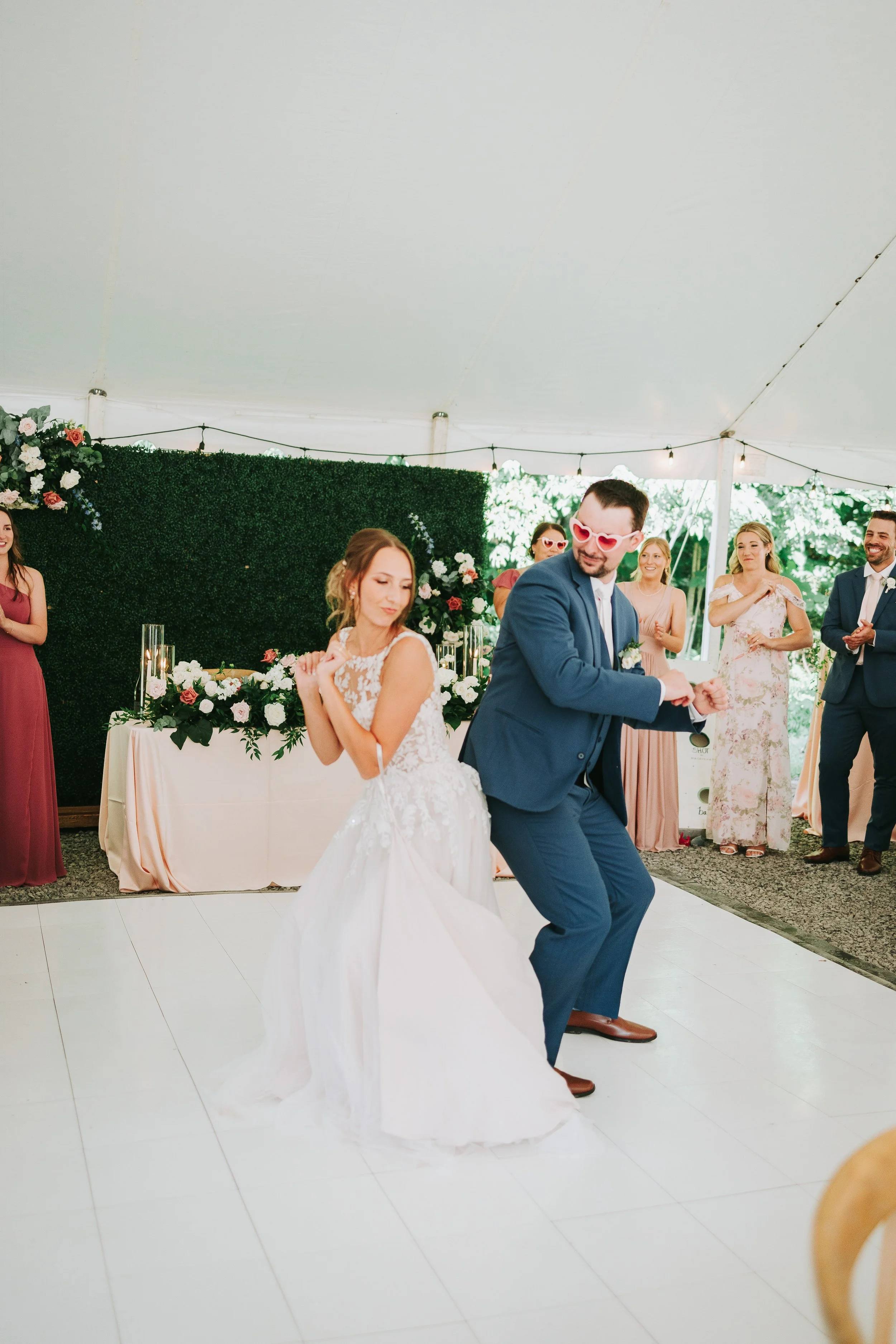 Bride and groom dancing at wedding reception inside a decorated tent, with guests watching and smiling in the background.