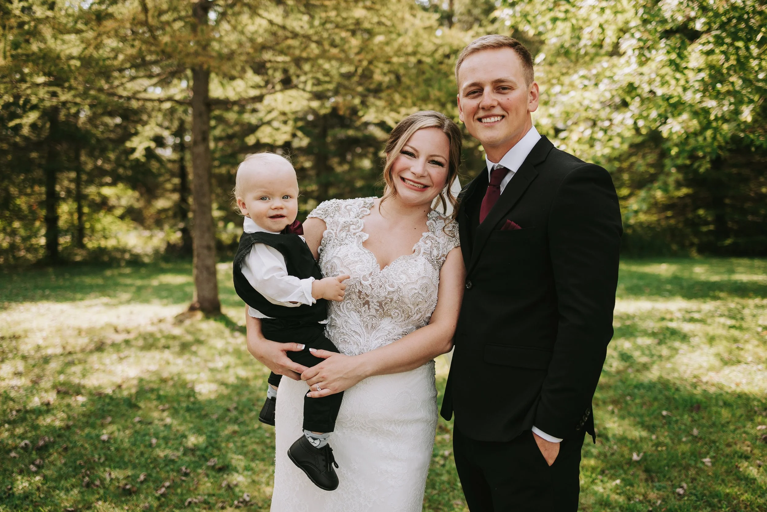 A bride in a white lace wedding dress, a groom in a black suit, and a young child in a tuxedo posing outdoors in a park with trees and green grass.