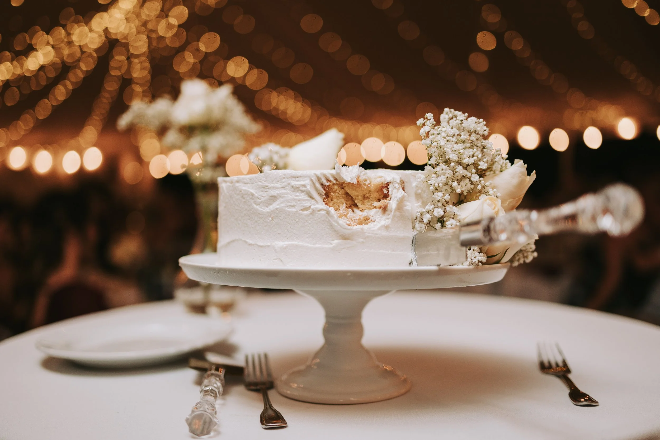 A partially eaten white wedding cake with white flowers on a cake stand. The background features blurred warm string lights.