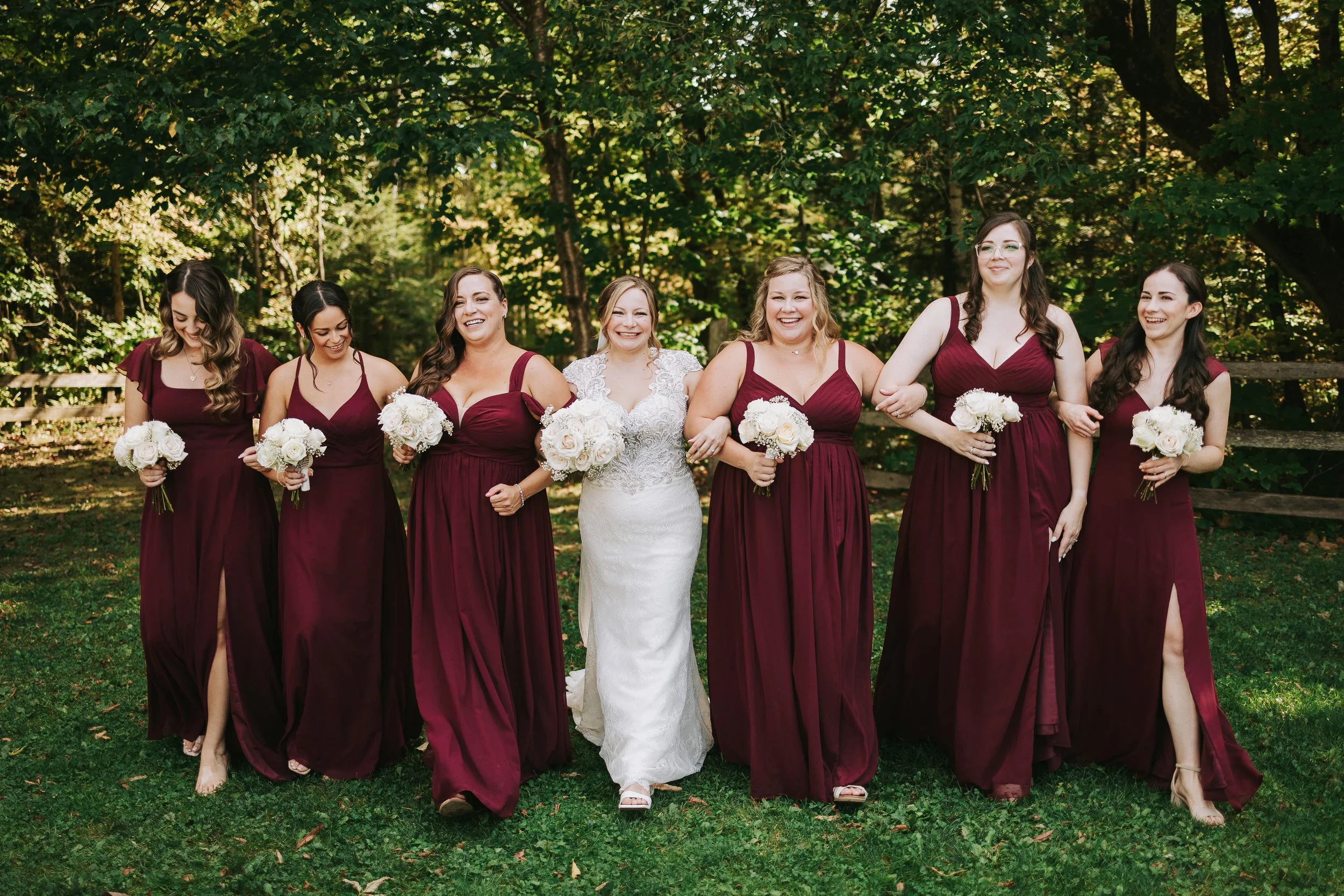 A bride and six bridesmaids walking outdoors on grass, smiling and holding white bouquets, with trees and a wooden fence in the background.