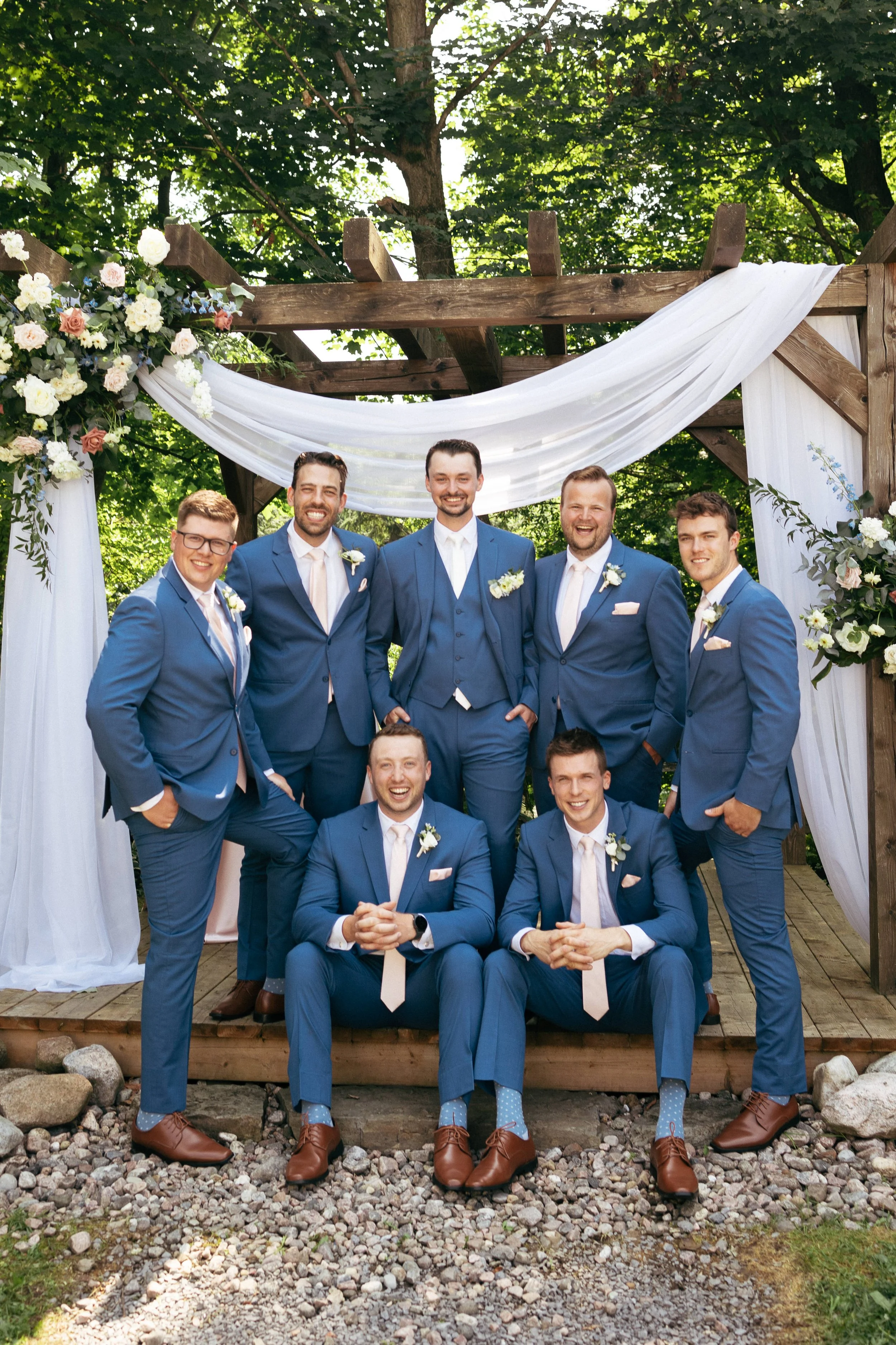 A group of eight groomsmen in matching blue suits with white shirts and ties, posing together at an outdoor wedding ceremony under a decorated wooden arch with white floral arrangements and drapery.