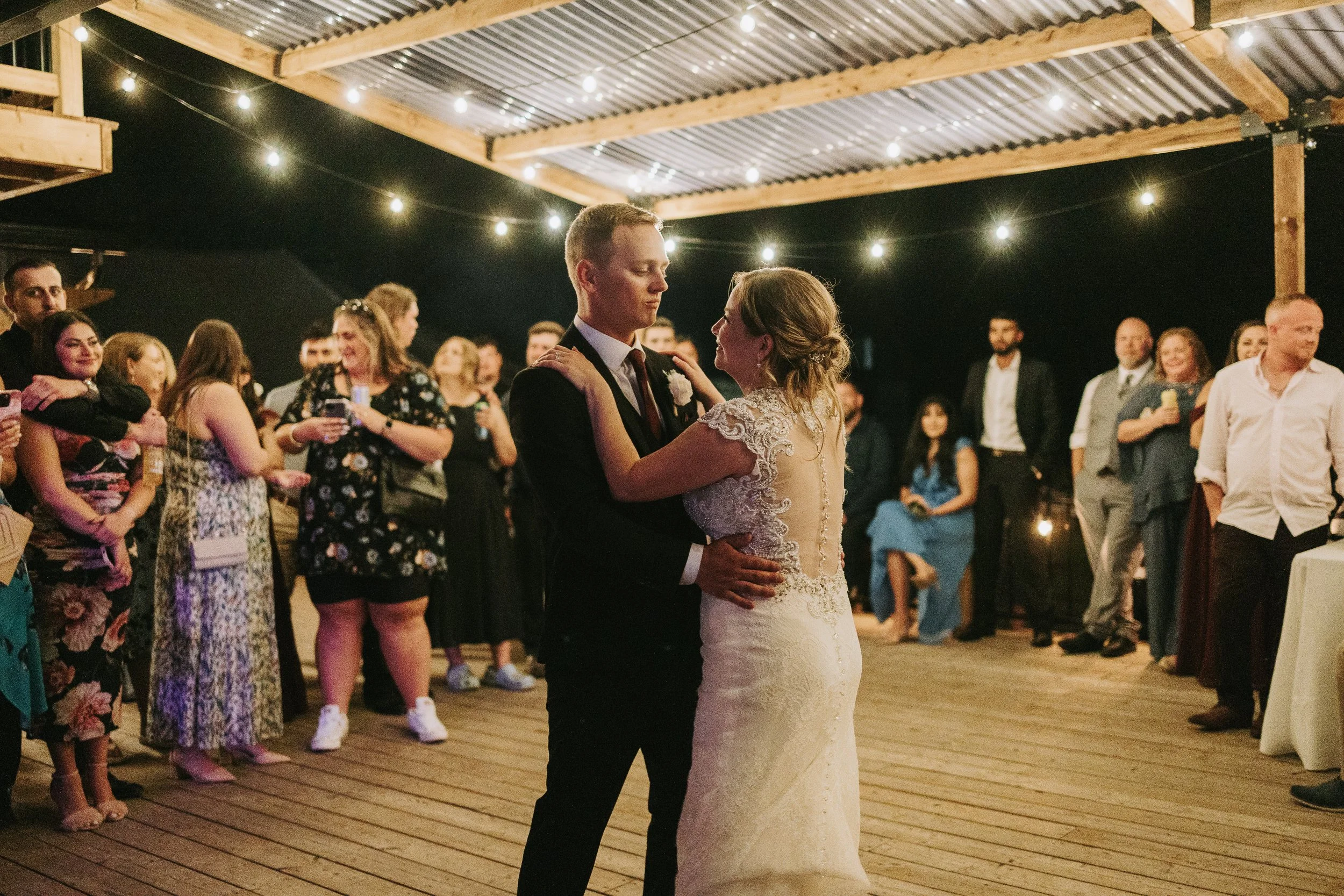 Couple dances during their wedding reception, surrounded by guests under string lights at a rustic venue.