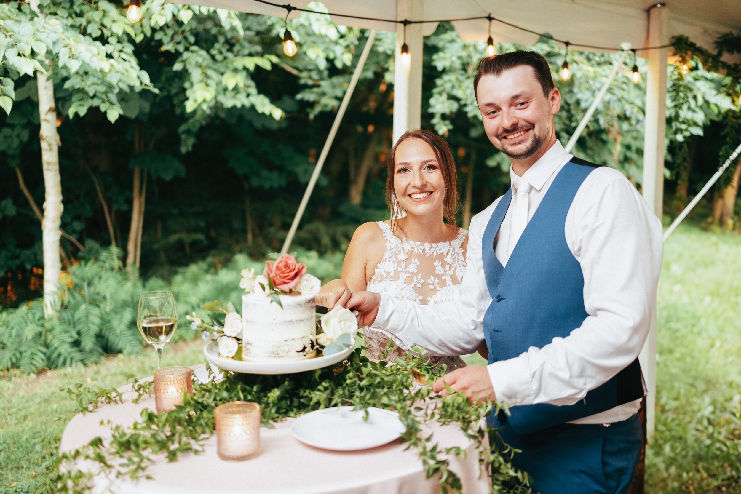 A smiling bride and groom cutting a wedding cake at an outdoor reception. The bride is in a lace wedding dress, and the groom is in a blue vest and white shirt. There are candles, a glass of white wine, and green foliage on the table, with string lig