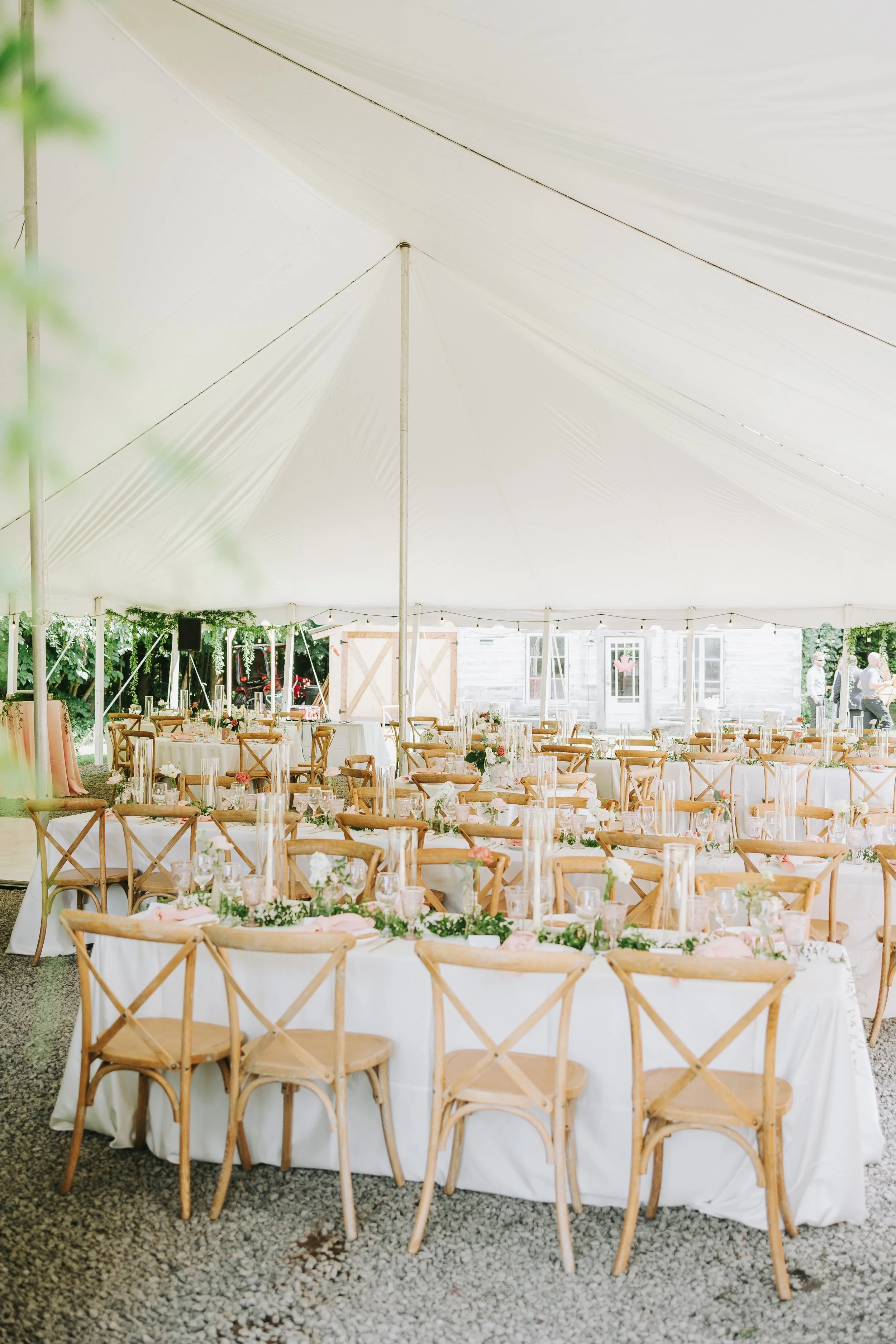 Decorated outdoor wedding reception under a large white tent with round tables and wooden chairs, floral centerpieces, and glassware.