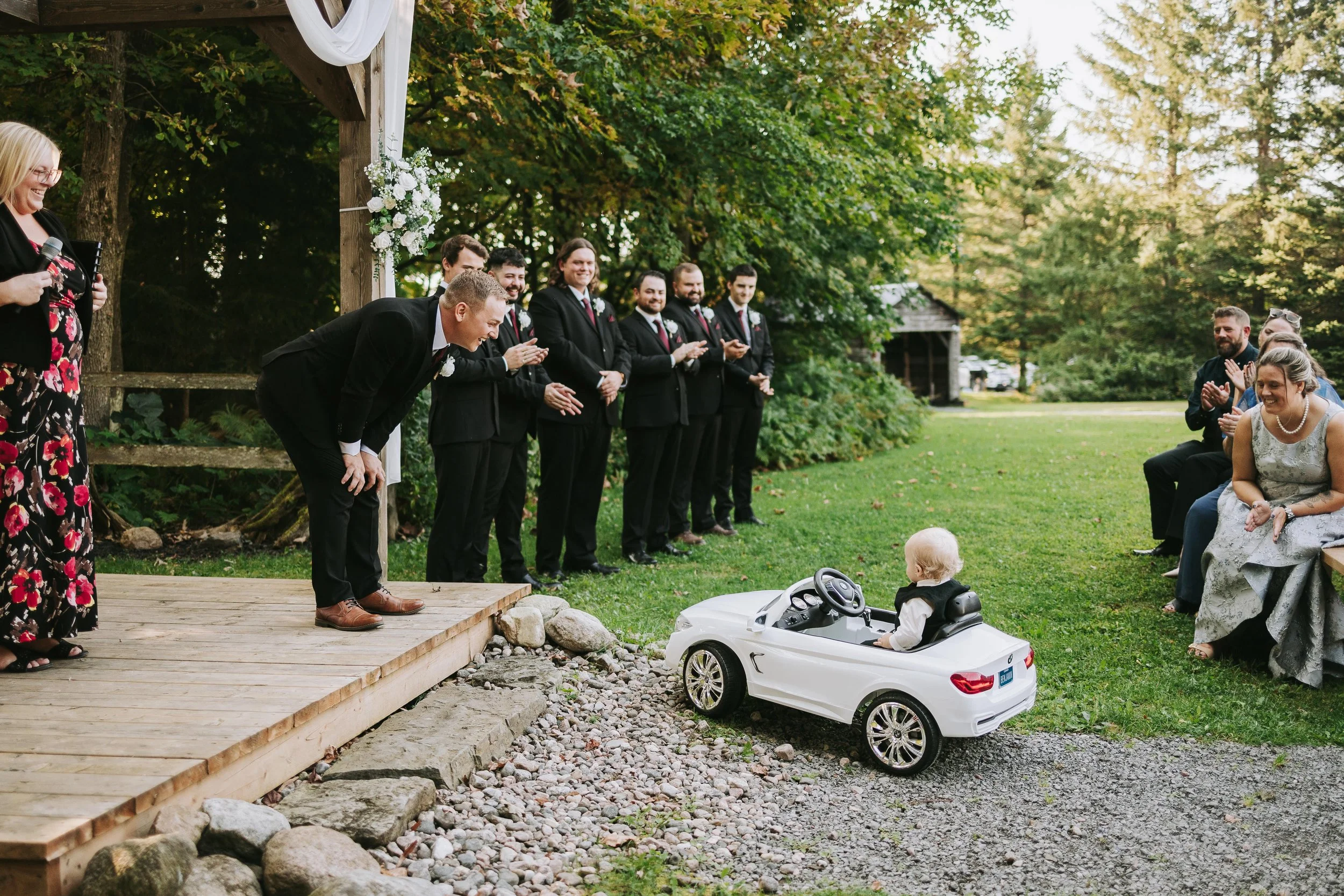A wedding ceremony outdoors with a child in a toy car approaching the bride and groom. The bride is sitting on a bench, and the groom is bowing on a small wooden stage near a group of groomsmen. Guests are seated, clapping and smiling, with trees and