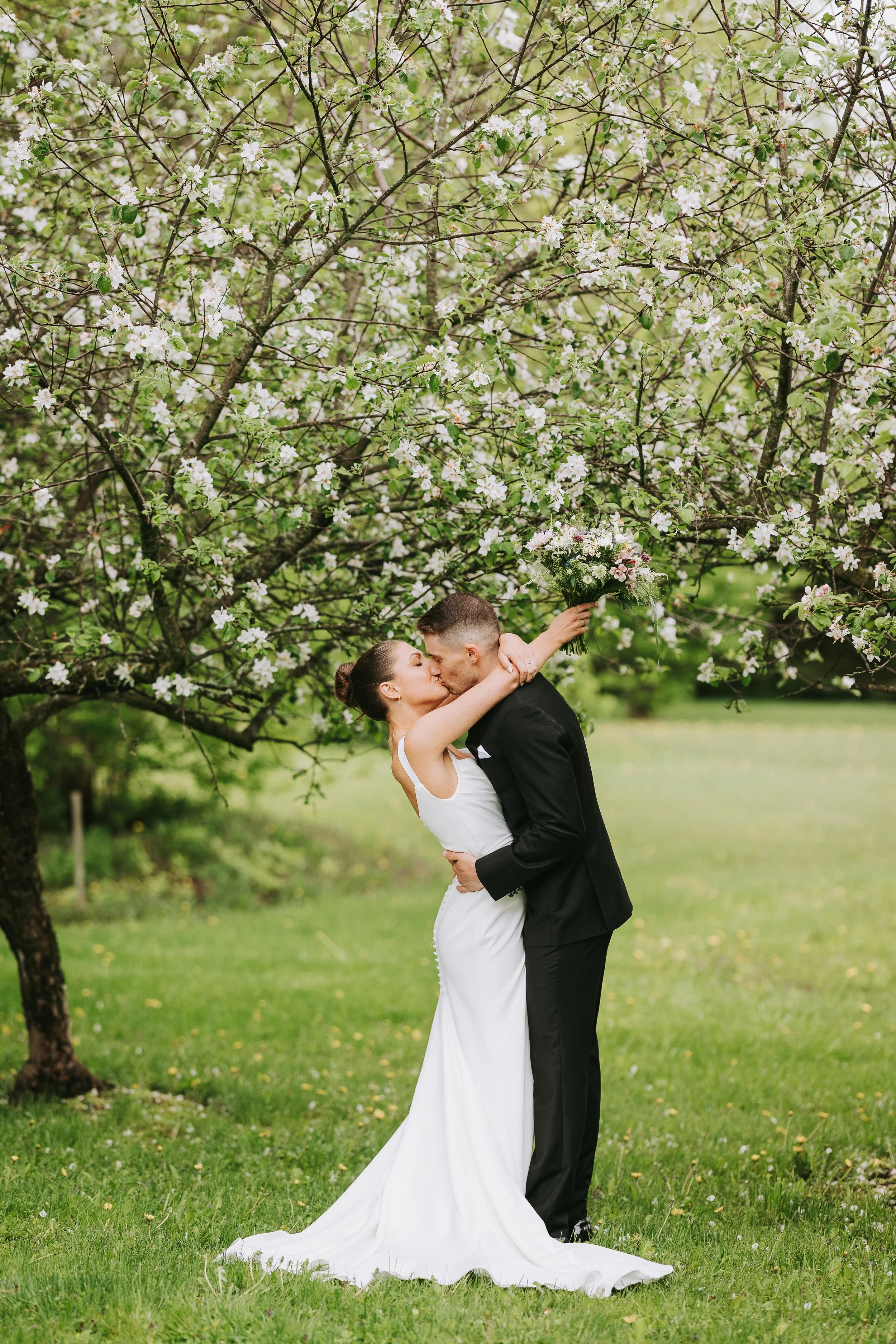 Bride and groom kissing under blooming tree on green lawn with bouquet in outdoor wedding setting.