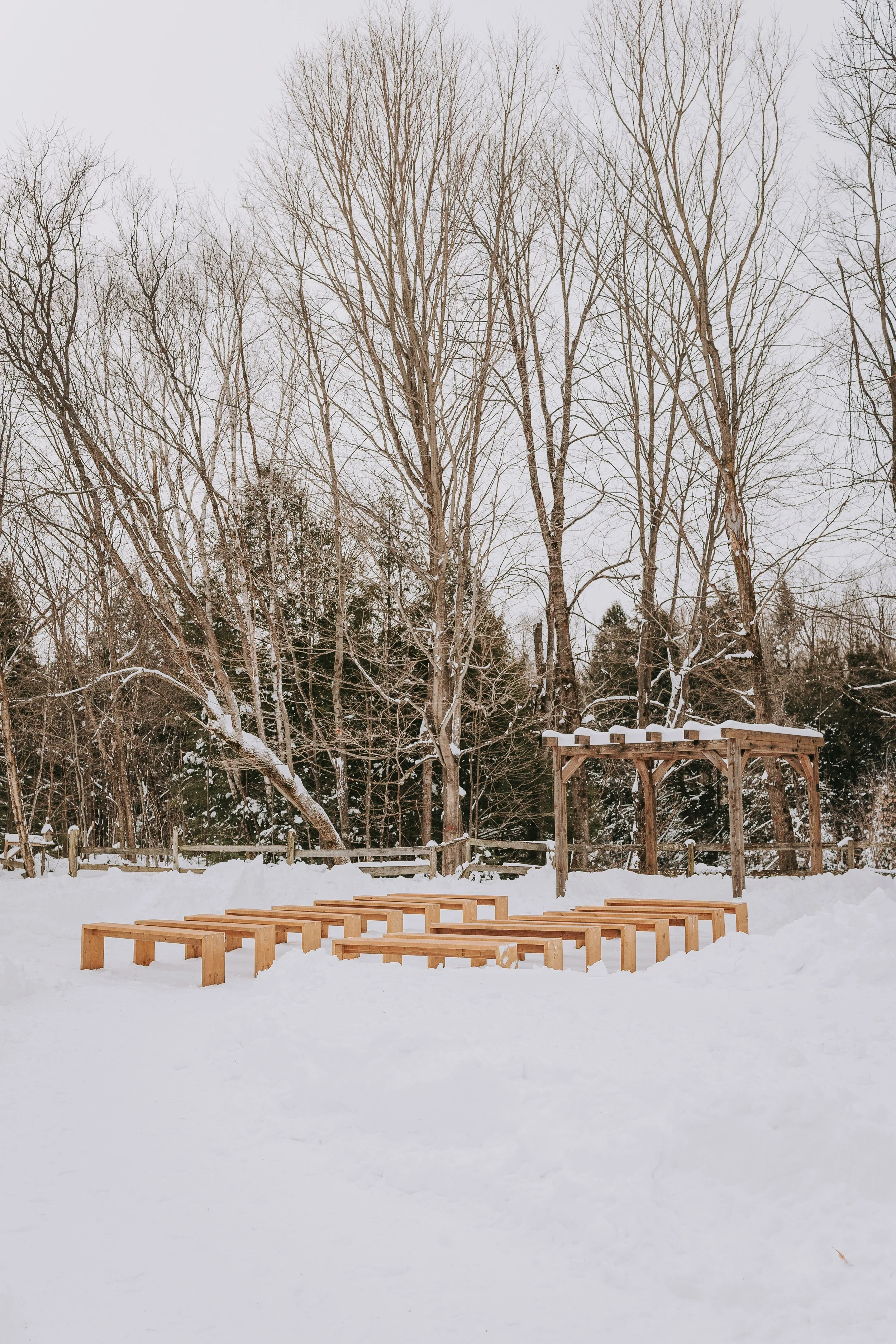 Snow-covered outdoor wooden structure with a raised platform, benches, and a trellis in a winter landscape with leafless trees in the background.