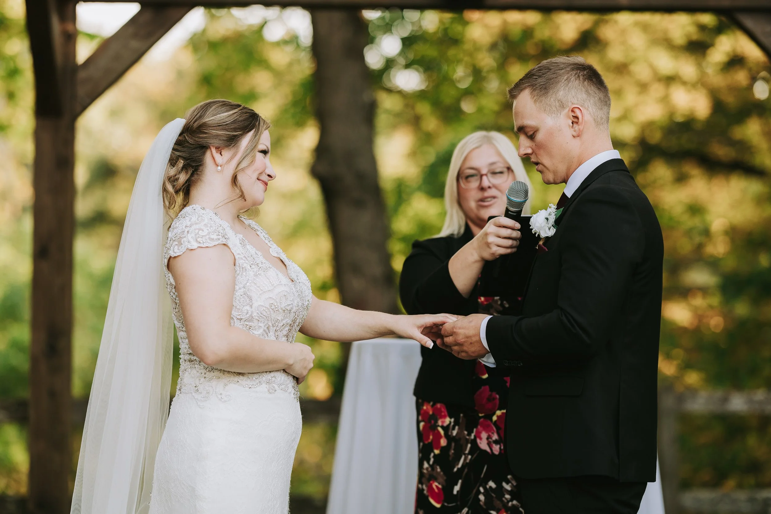 A bride and groom exchanging rings during their wedding ceremony outdoors, with a officiant holding a microphone in the background.