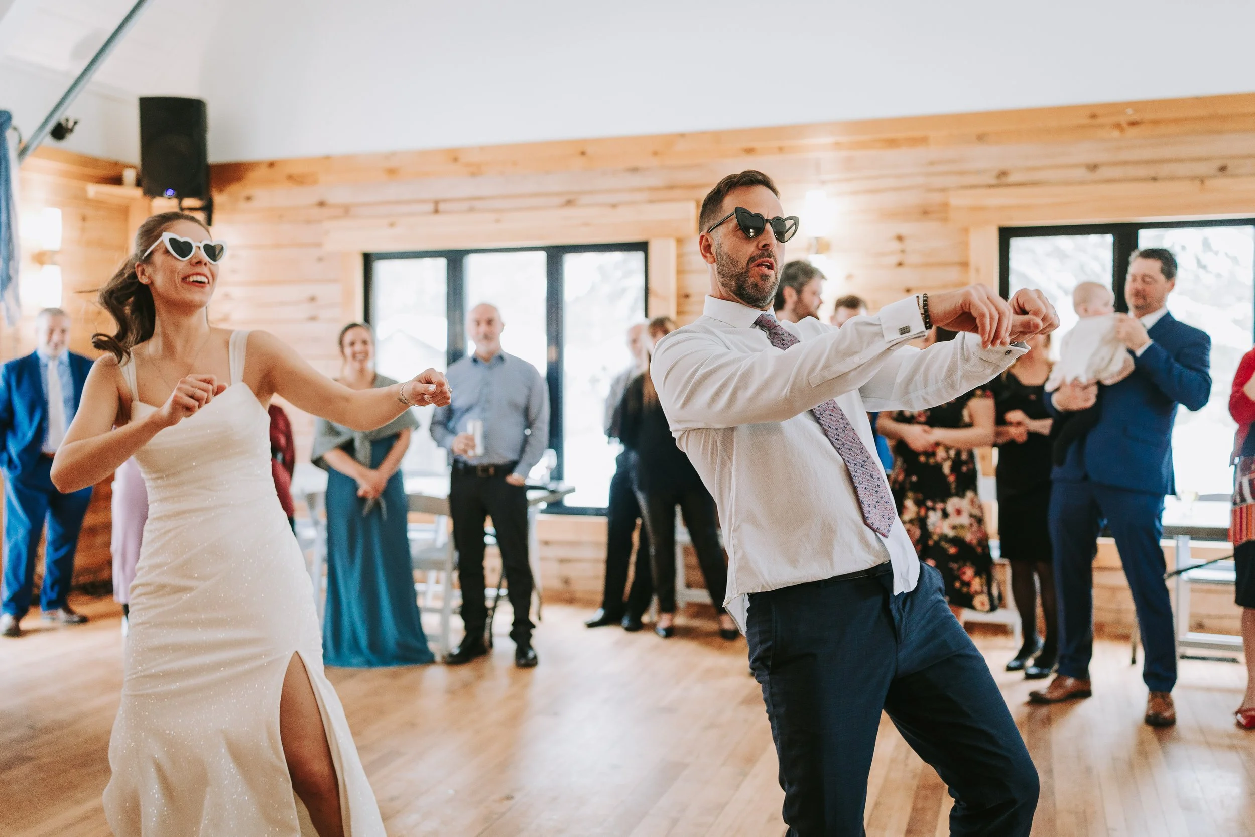 People dancing at a celebration in a wooden indoor venue with large windows and a lively atmosphere.