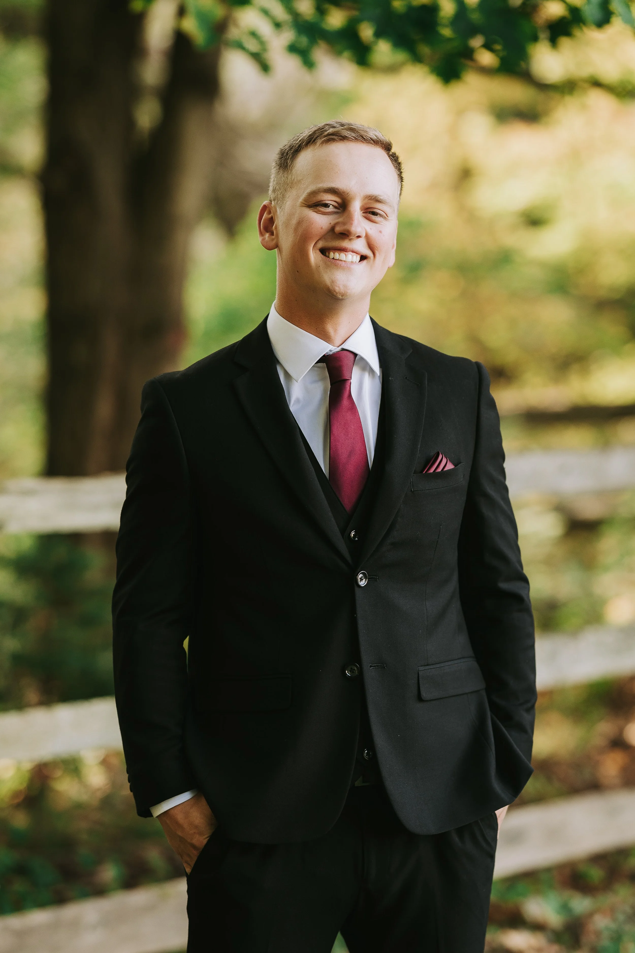 A young man in a black suit with a white shirt and maroon tie standing outdoors near a wooden fence with greenery and trees in the background, smiling at the camera.