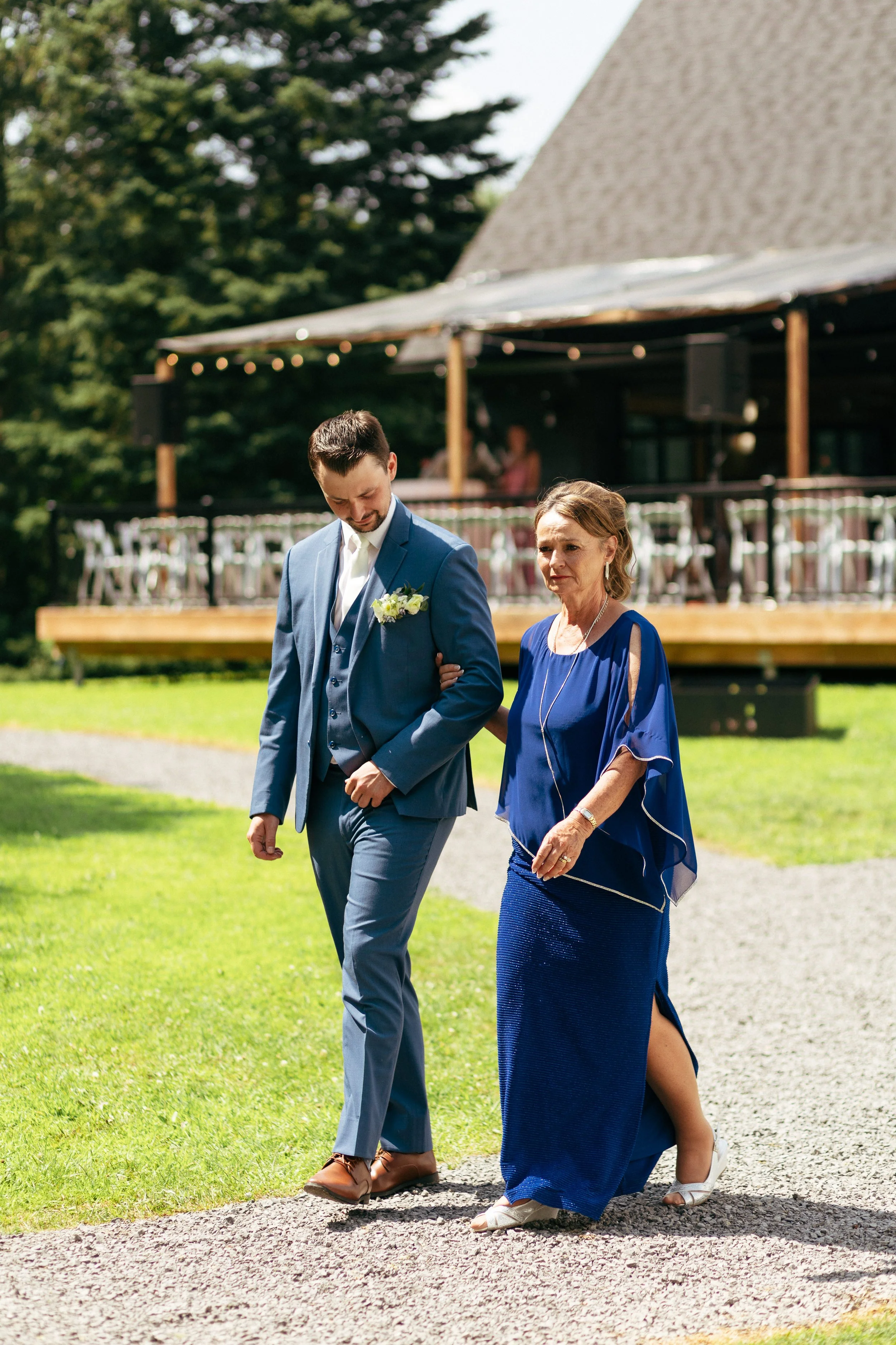 A groom walking arm-in-arm with an older woman outdoors during a wedding, with a wooden building and string lights in the background.