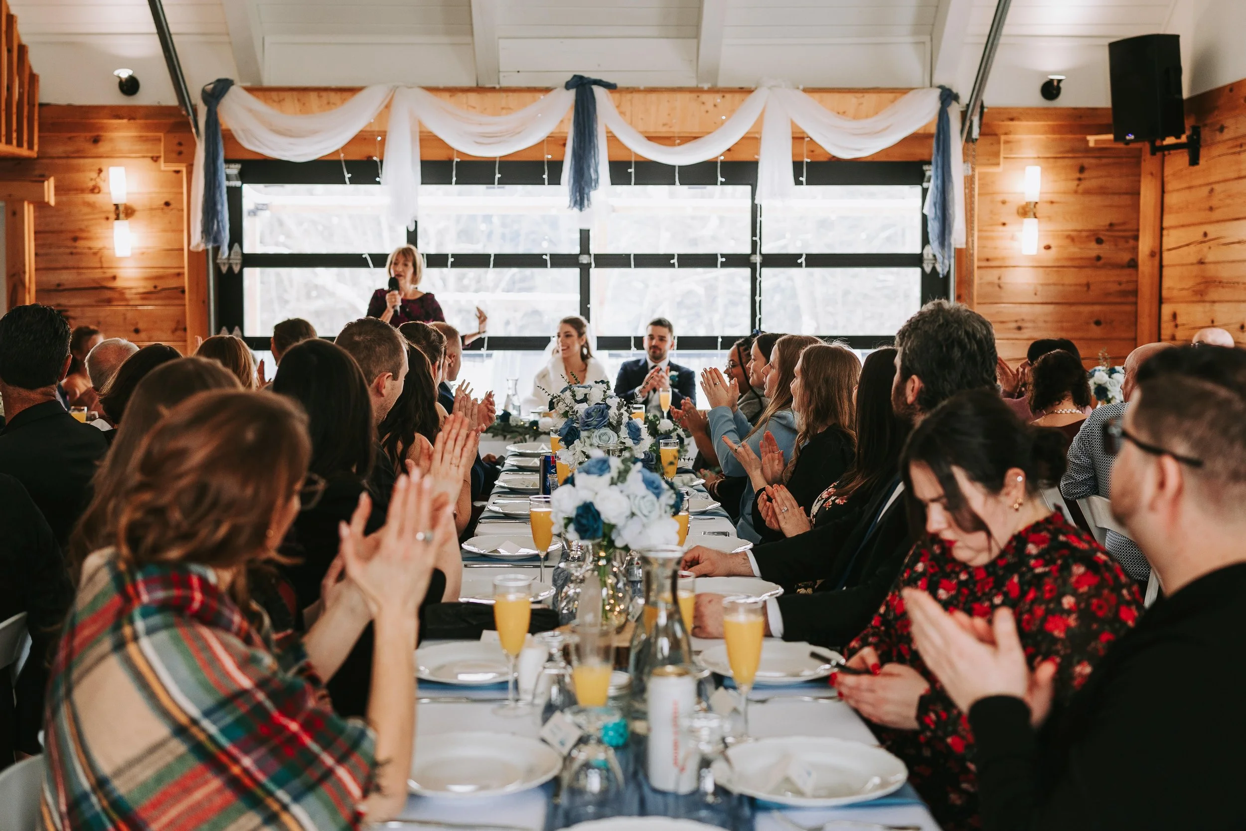 Guests at a wedding reception seated at a long table, clapping as a woman gives a speech. The table is decorated with flowers and drinks. The background features large windows and rustic wooden walls.