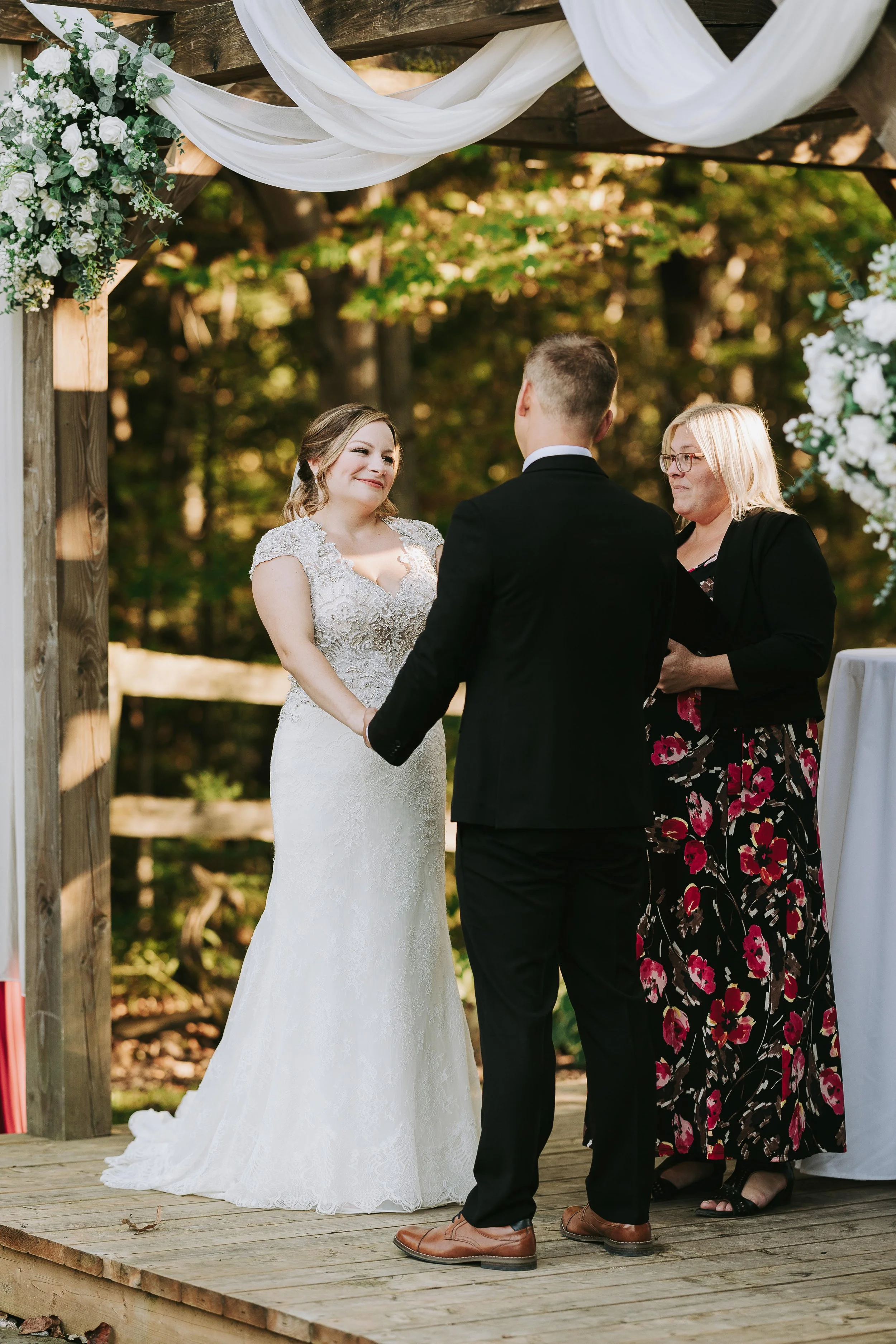 A couple getting married outdoors, holding hands and exchanging vows under a decorated wooden arch with white and green floral arrangements and drapery.