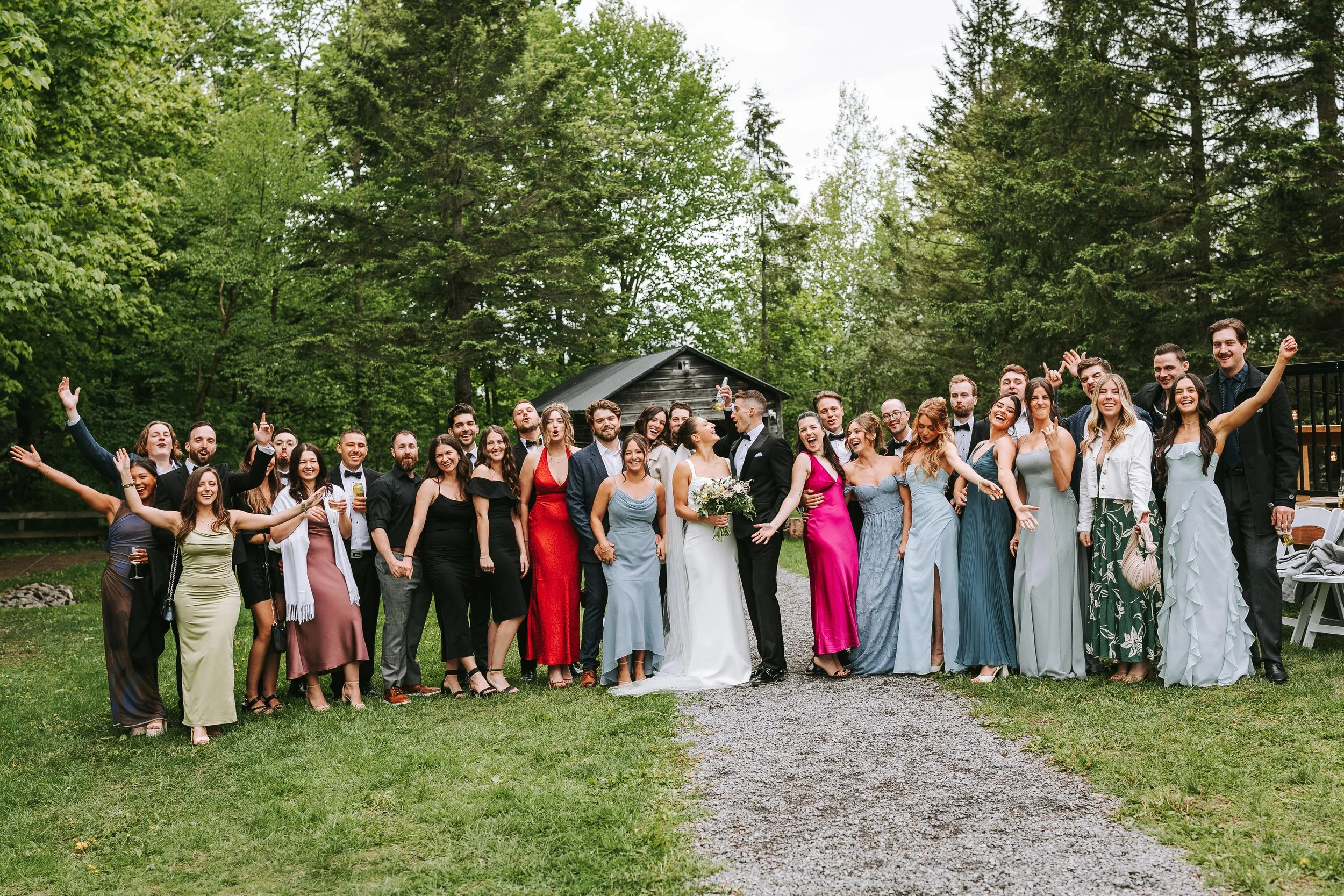 Group of happy people in formal attire at a wedding, outdoors on a grassy area with trees and a wooden cabin in the background.
