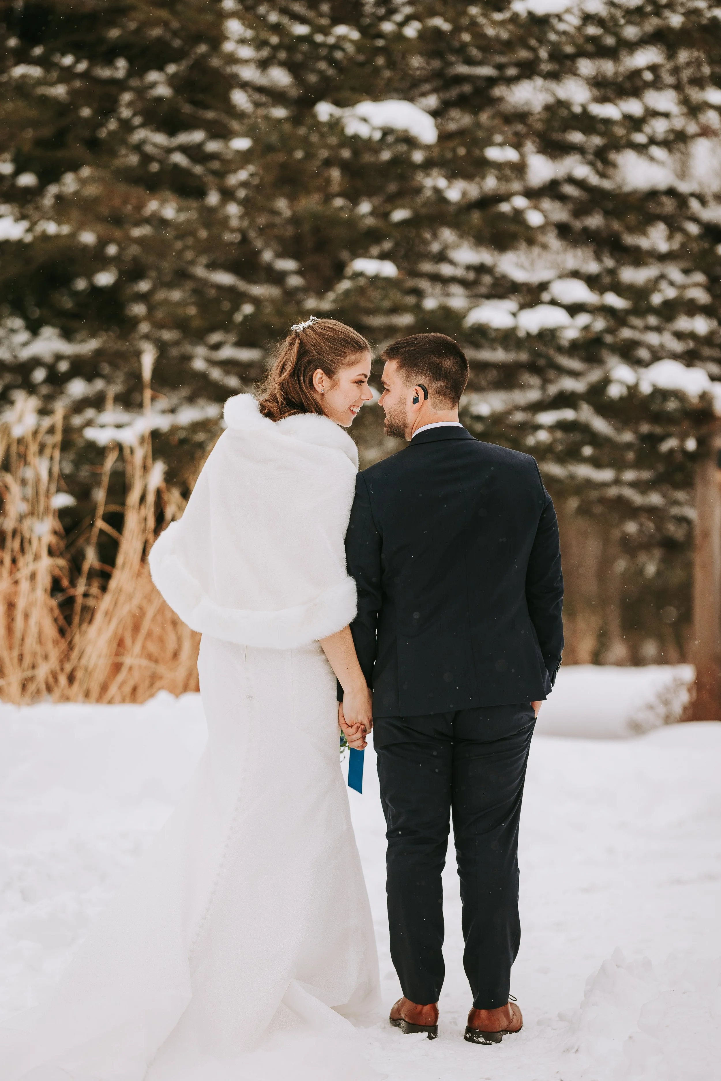 A bride and groom holding hands and standing close together outdoors in a snowy forest, gazing at each other before their wedding ceremony.