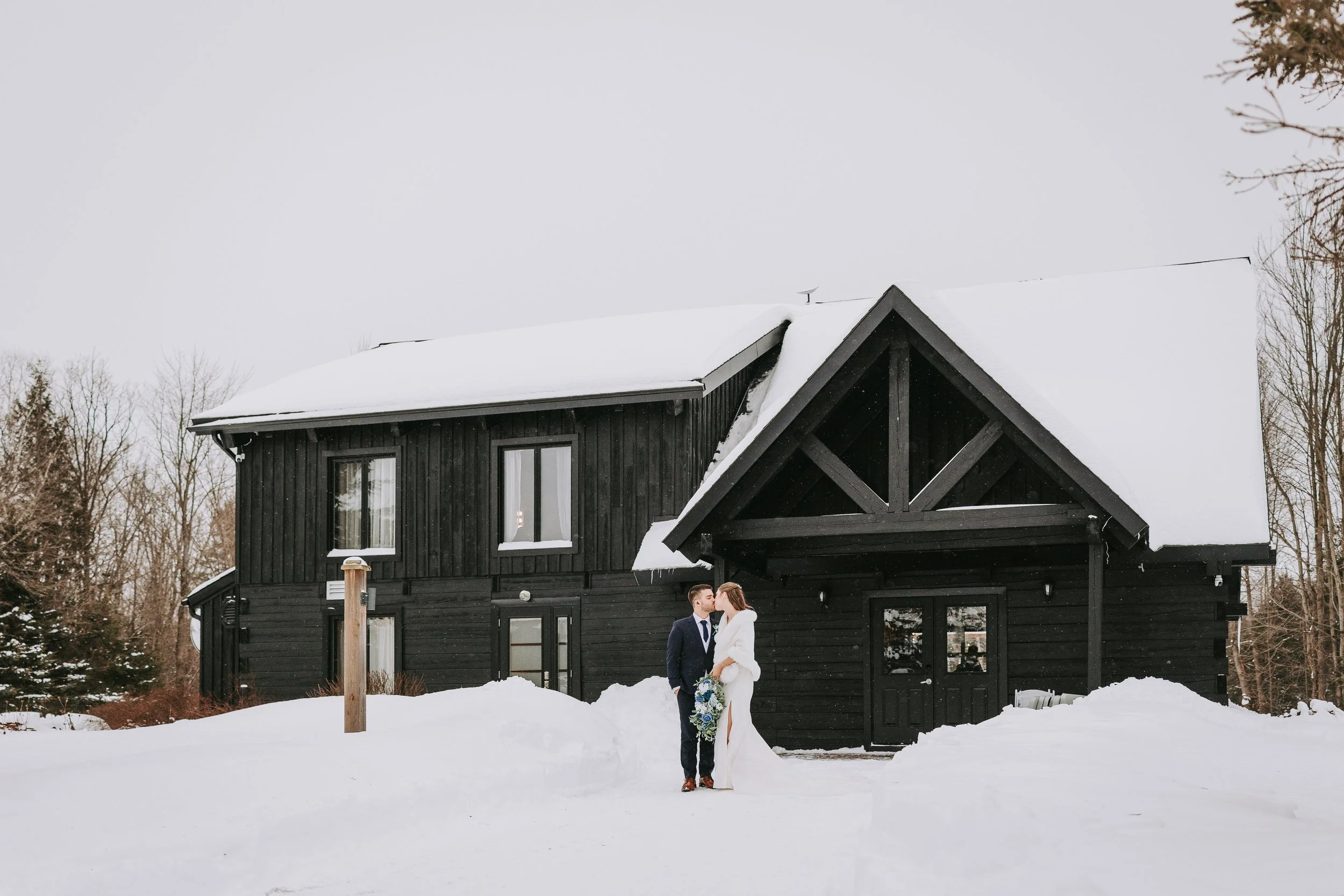 A newlywed couple stands in the snow in front of a black wooden venue with a snow-covered roof, surrounded by leafless trees on a winter day.