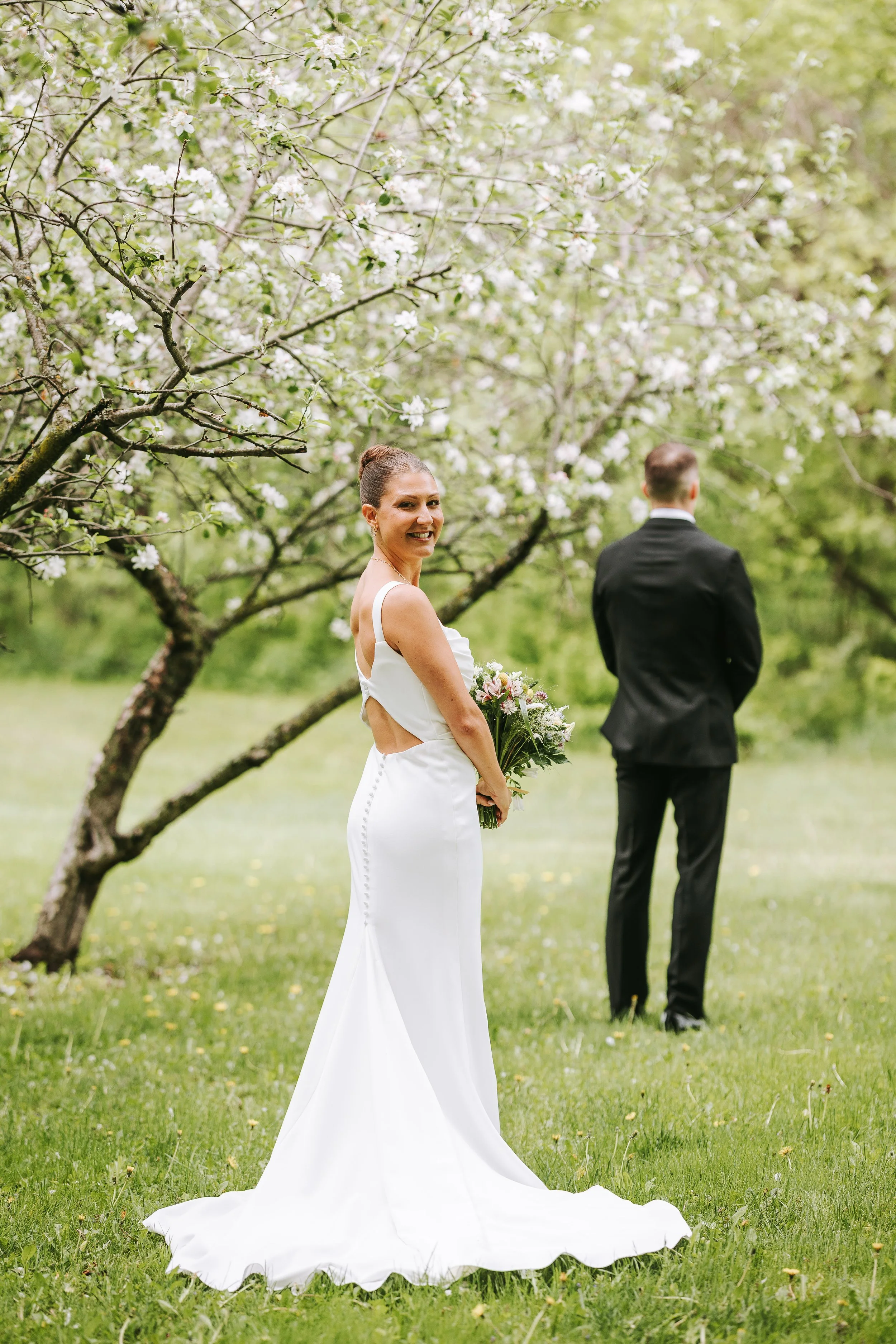A bride in a white wedding dress smiling and holding a bouquet, standing outdoors under a blooming tree, with a groom in a black suit standing in the background facing away.