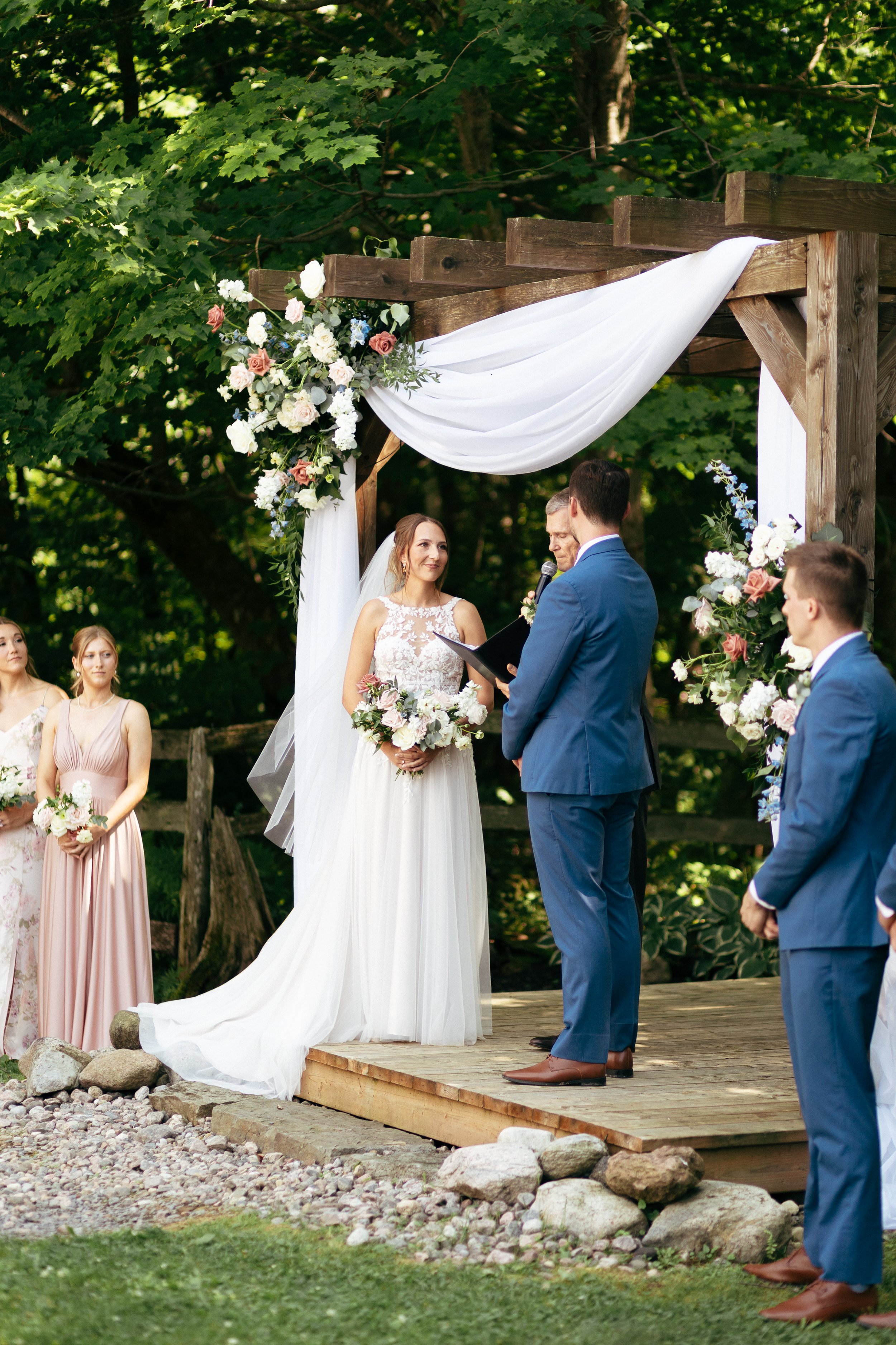 A bride and groom exchange vows during a wedding ceremony outdoors on a wooden stage decorated with flowers and white fabric, with bridesmaids standing beside them.