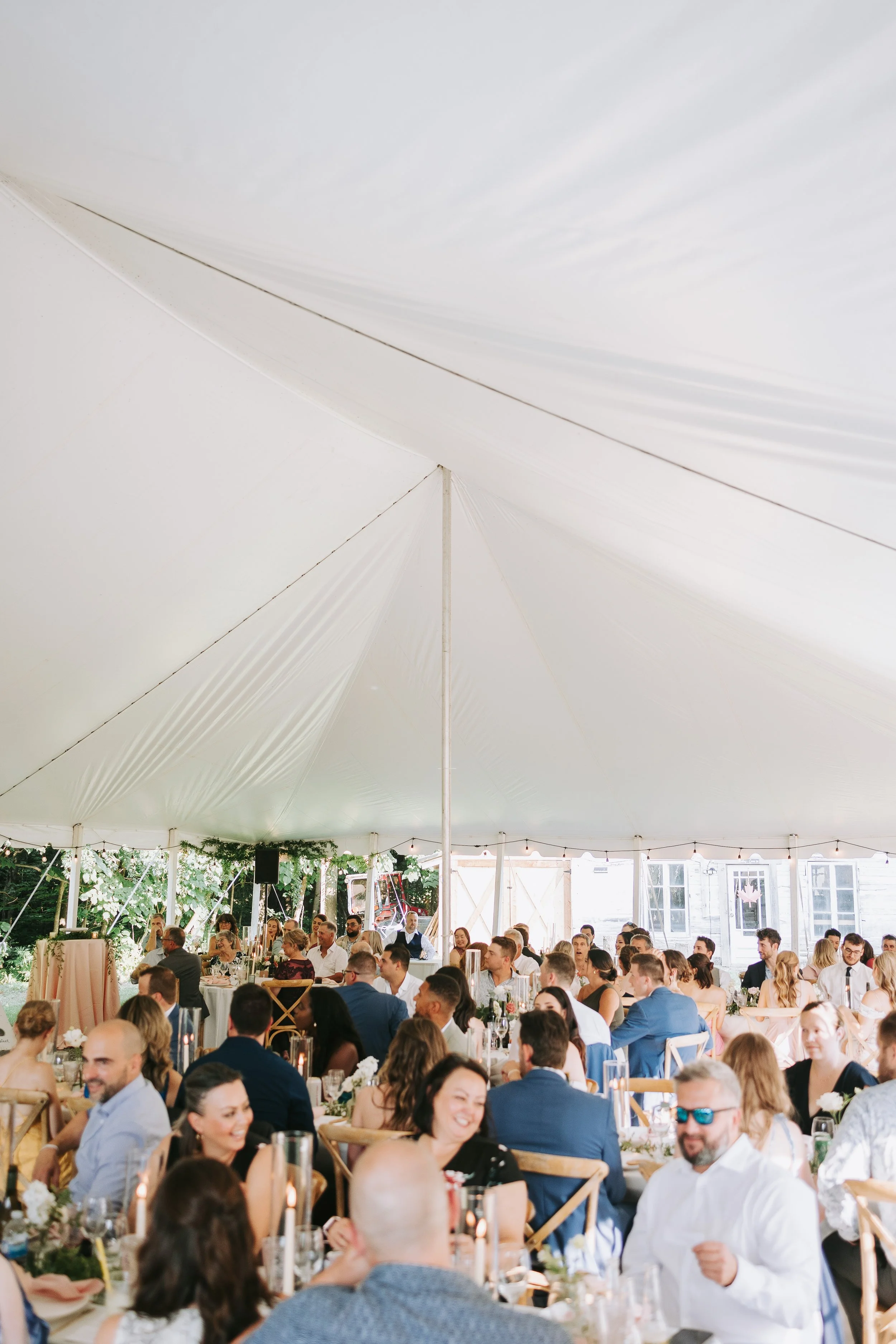 People gathered under a large white tent at a celebration or wedding, sitting at decorated tables with floral centerpieces.